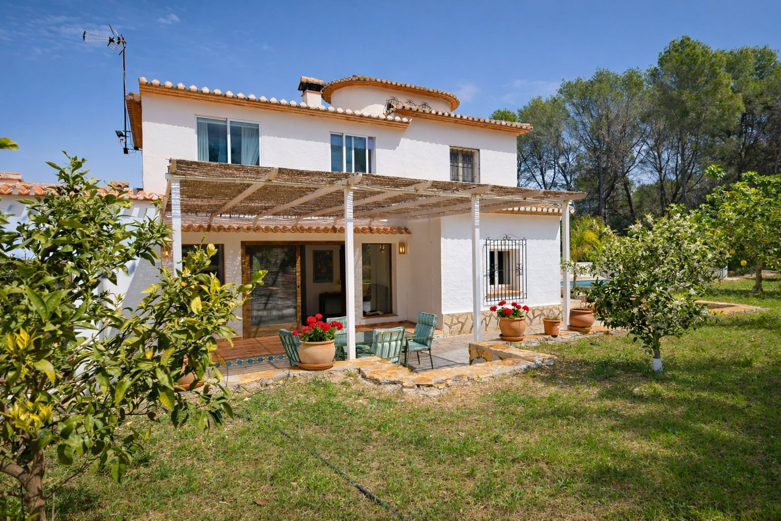 Rear facade of the villa featuring a reed-covered porch, outdoor dining area, and a garden with fruit trees. The Mediterranean design includes stone accents and terracotta tiles.