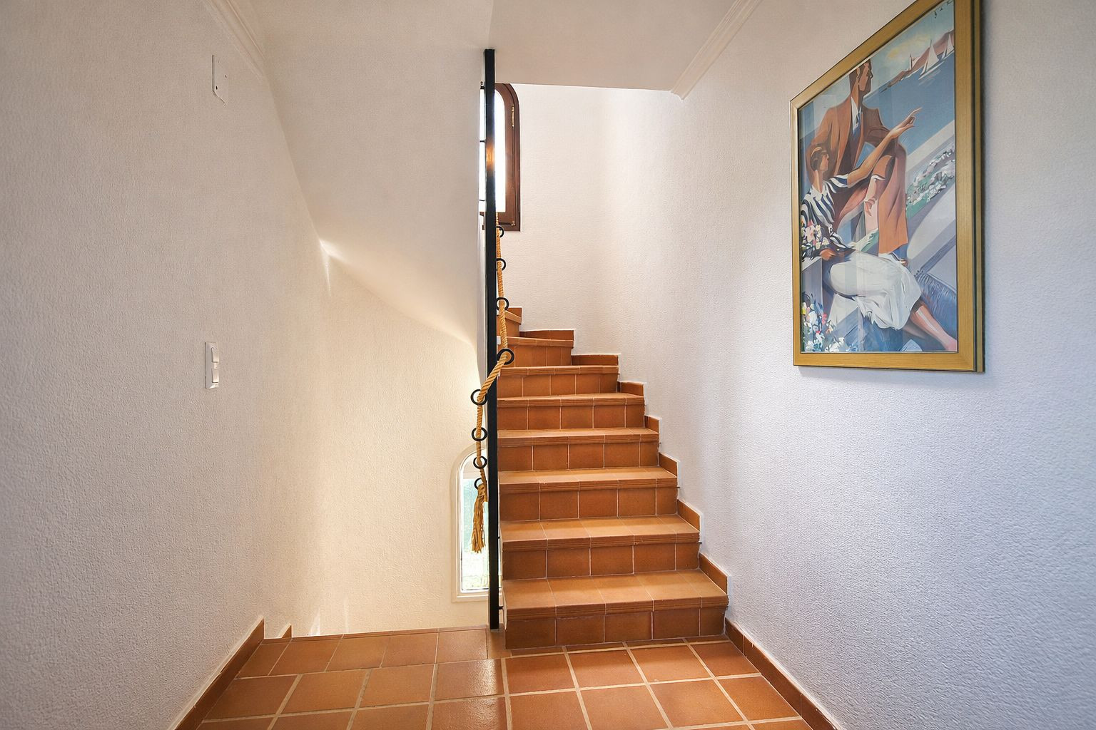 Terracotta tiled staircase featuring a black metal handrail with rope detailing, set against white walls with natural light and wall art.