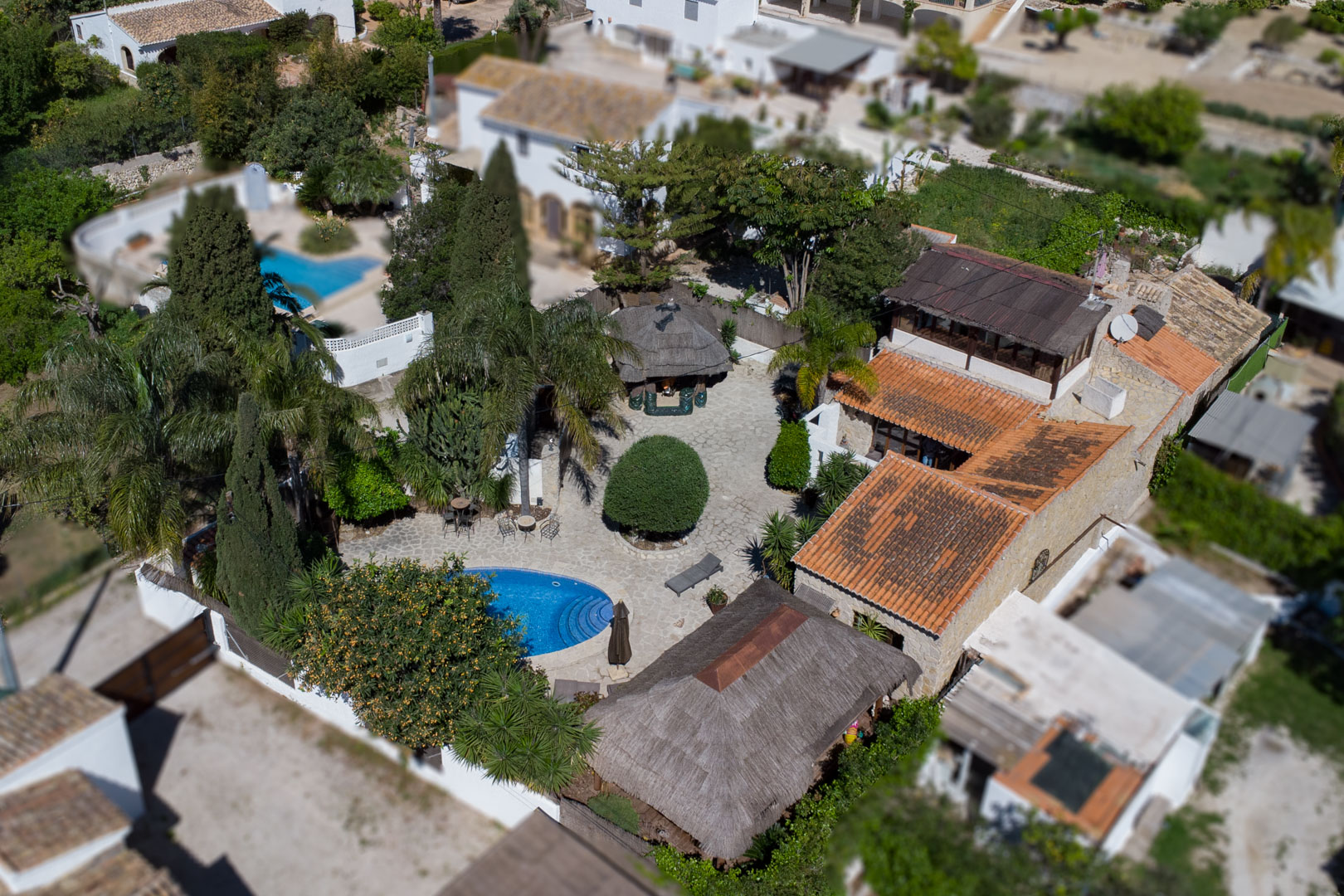Aerial view of the estate showing the oval pool, natural stone patio, and Balinese thatched pergolas surrounded by lush Mediterranean trees and stone walls.