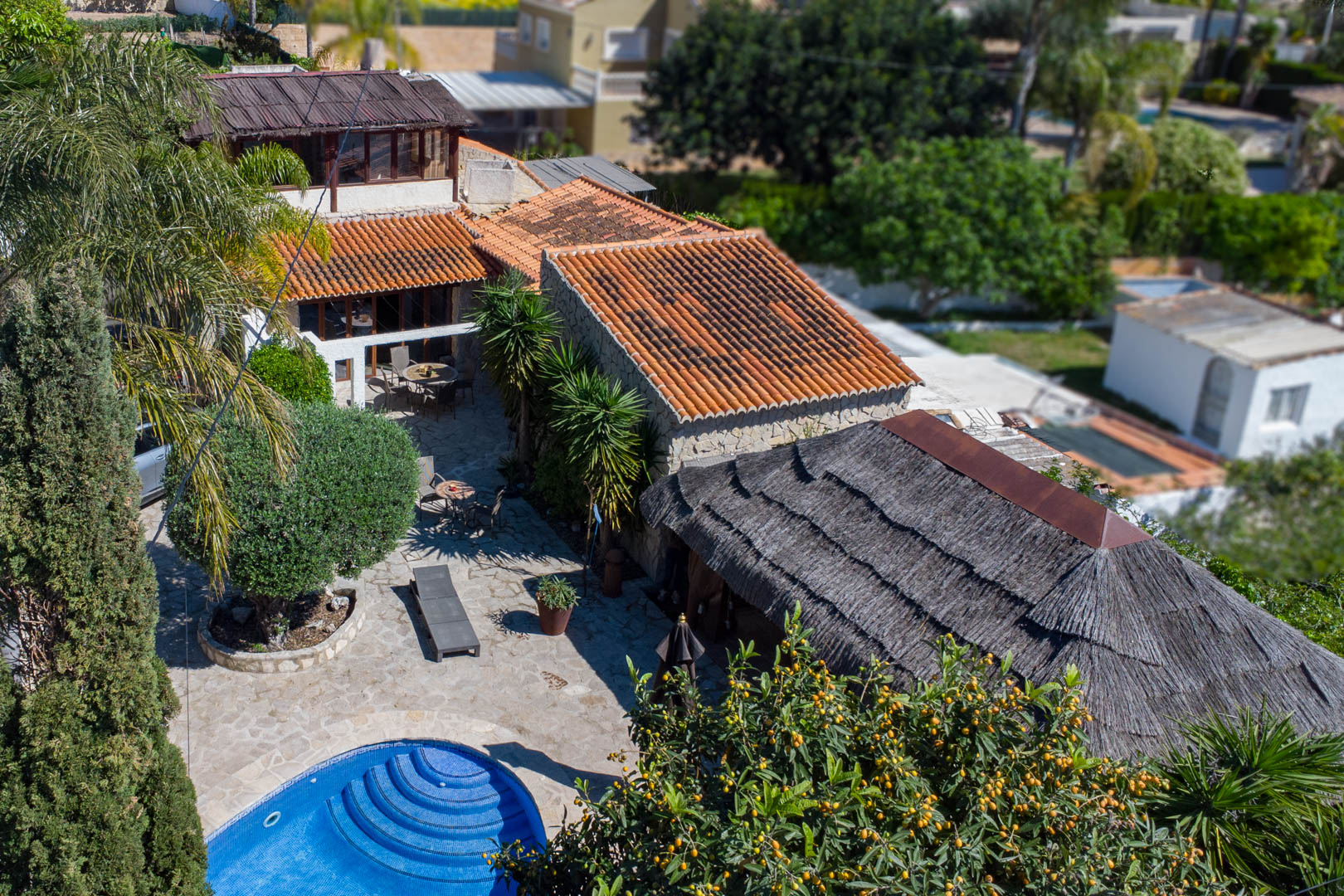 Aerial view of the rustic villa featuring terracotta roofs, a circular blue tiled pool, extensive stone terraces, and a large thatched Balinese pergola.