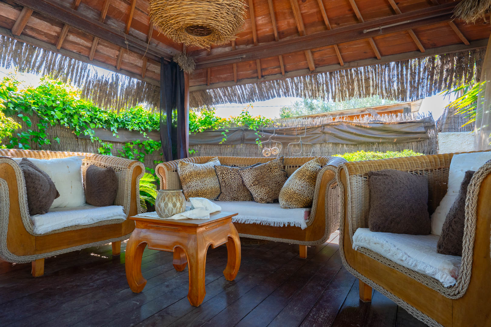 Balinese pergola featuring an exposed wooden ceiling, wicker lounge furniture with leopard print accents, and a rustic coffee table on dark wood decking.
