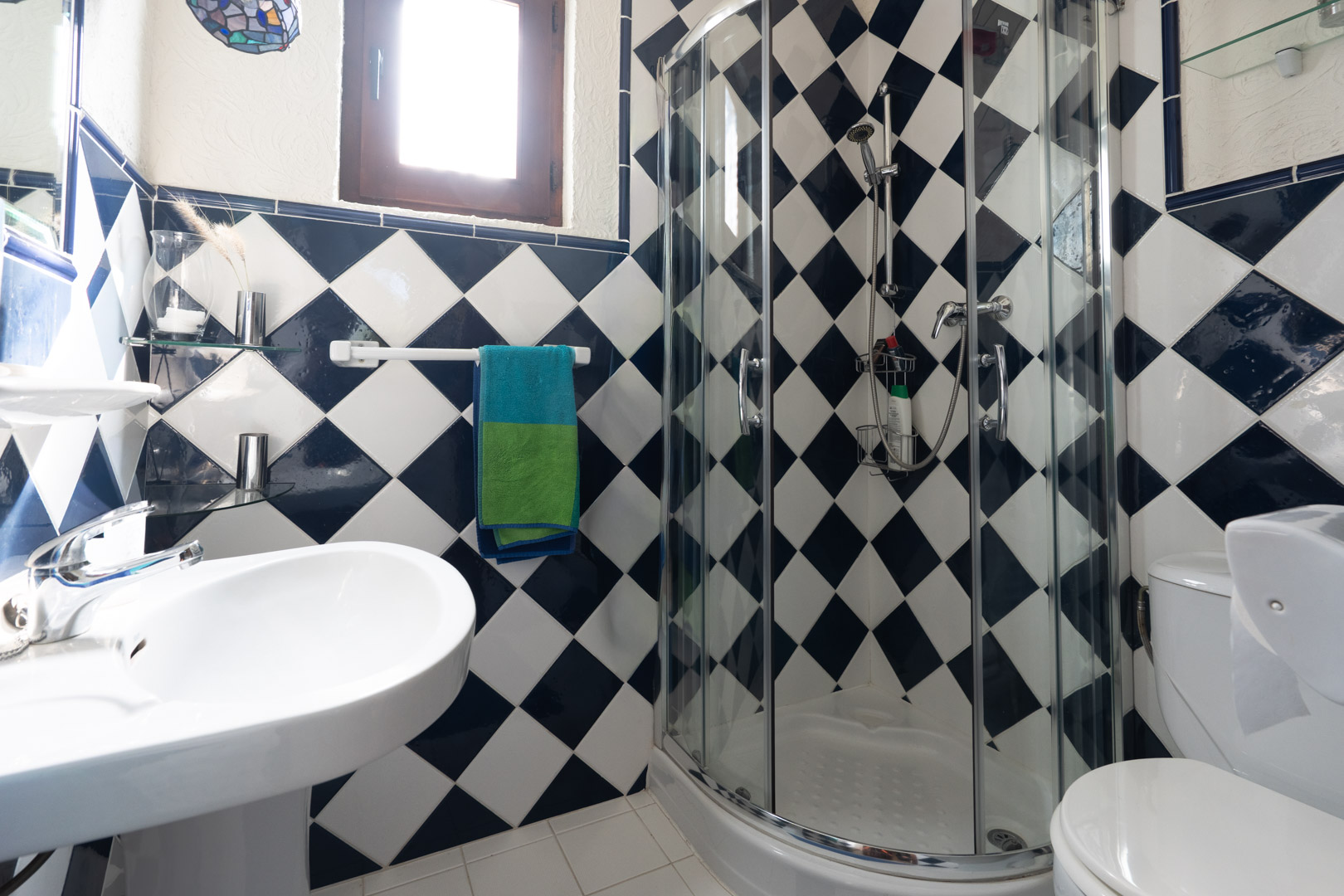 Bathroom featuring blue and white checkered wall tiles, a curved corner shower cabin with glass doors, and a white pedestal sink under natural light.