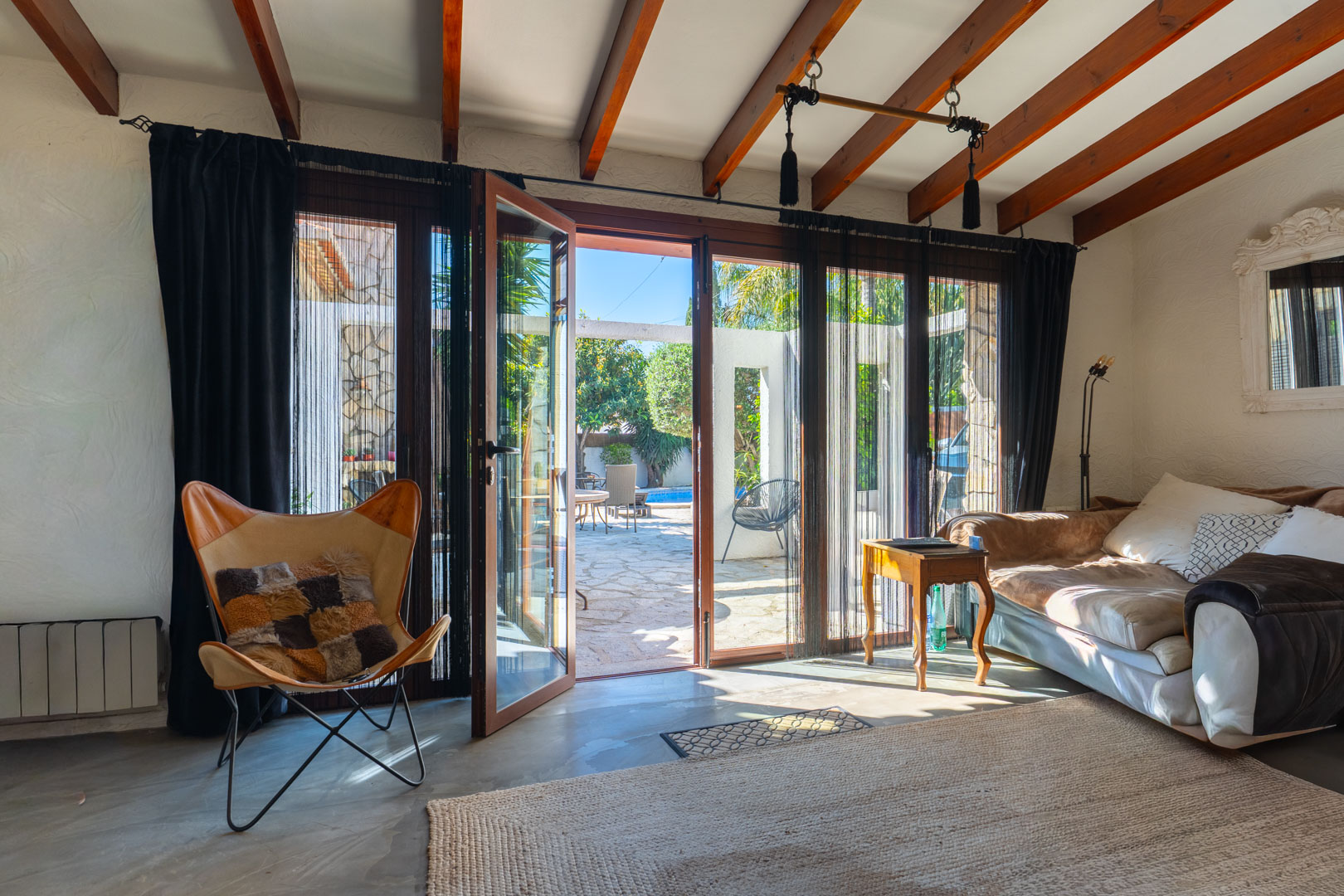 Bright living area featuring exposed wooden ceiling beams and large glass doors that open onto the stone-paved patio and pool area.