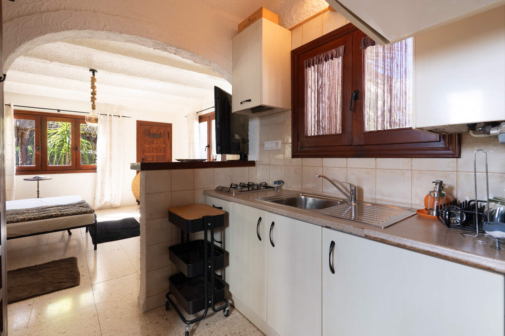 Compact rustic kitchen featuring white cabinetry and wooden window frames, connected to a bright bedroom through a Mediterranean archway.