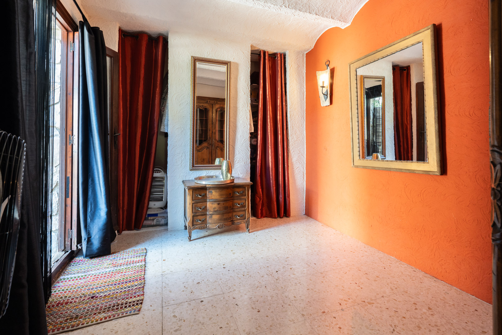 Dressing area featuring an orange accent wall, ornate mirrors, a classic wooden chest of drawers, and terrazzo flooring. Red curtains conceal the integrated closet spaces.