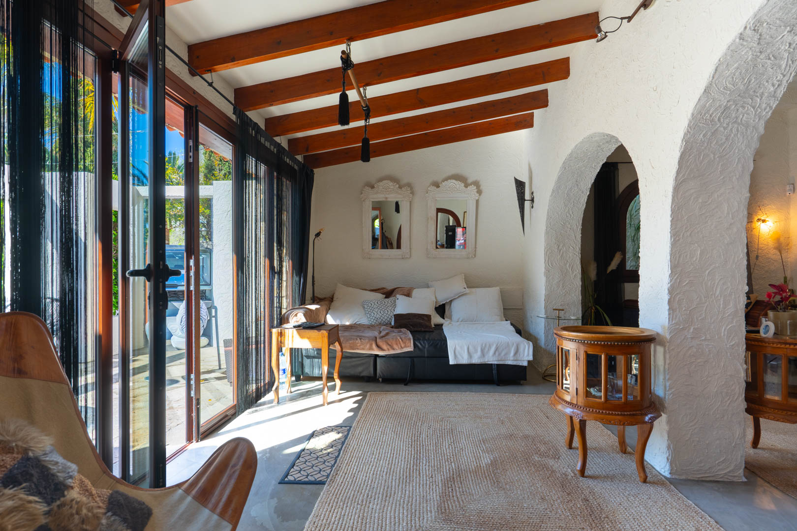 Glazed naya featuring exposed wooden ceiling beams, textured white walls, and a traditional archway. Large glass doors with string curtains provide abundant natural light to the lounge area.