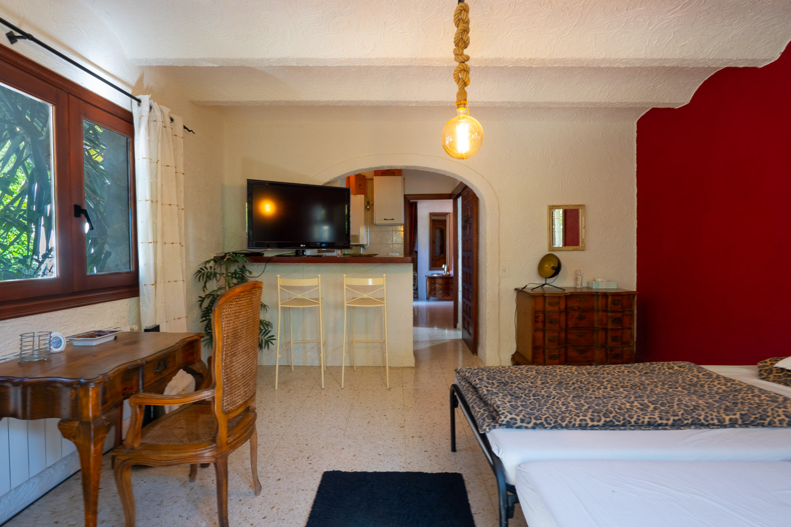 Guest bedroom featuring a classic wooden desk, red accent wall, and a breakfast bar with stools connecting to the kitchen through a traditional archway.