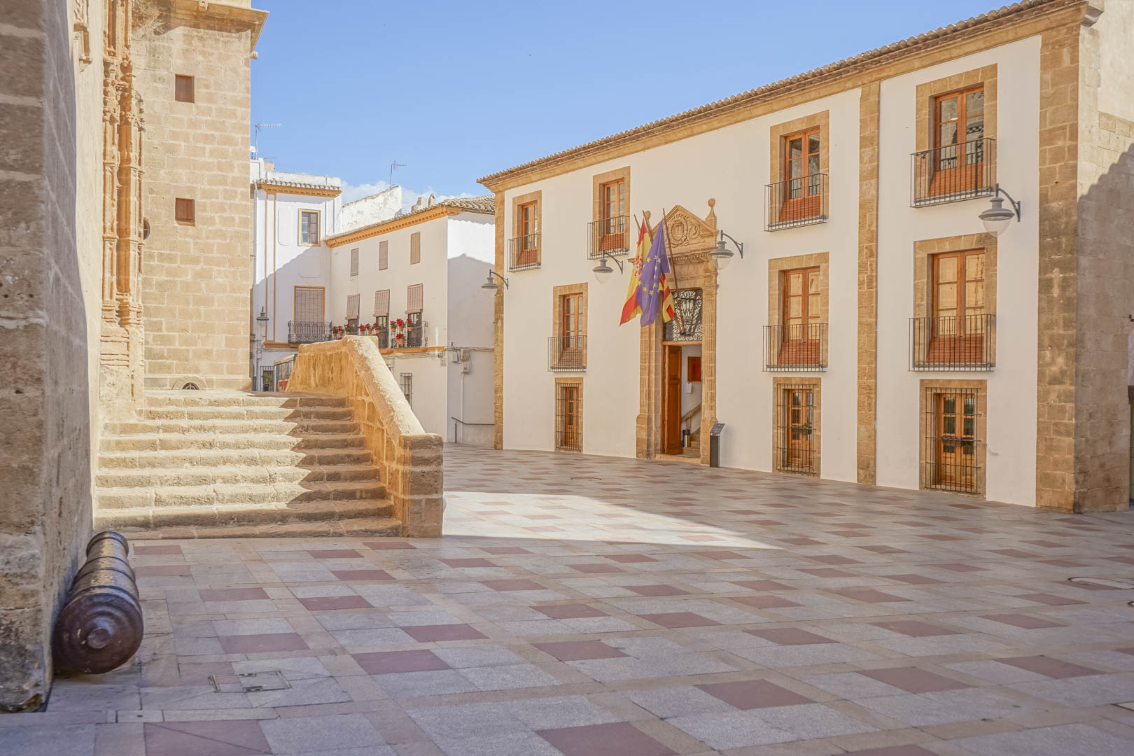 Historic town square in Jávea featuring traditional stone architecture, the local town hall with flags, and the fortified San Bartolomé church.