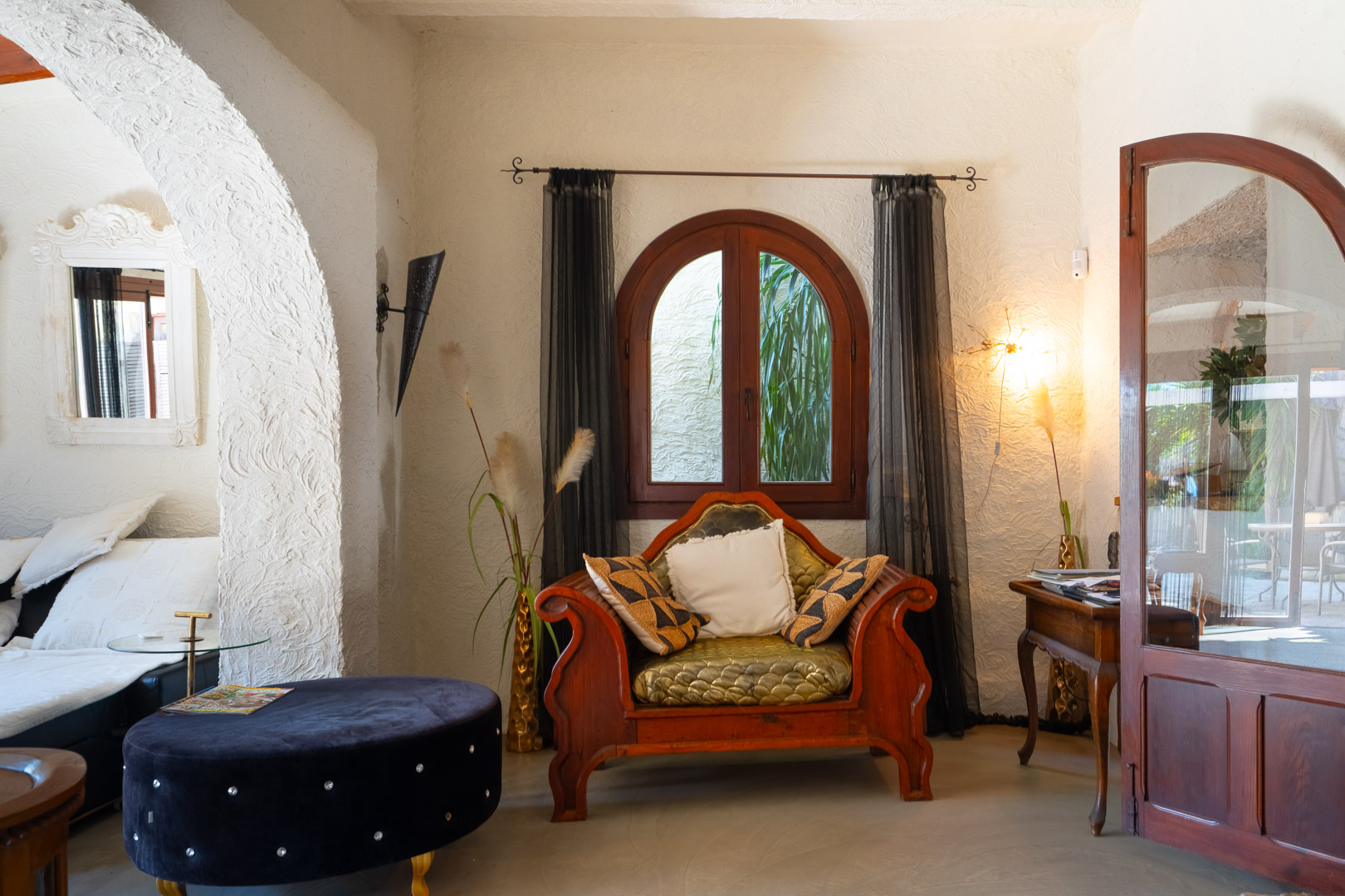 Living area featuring textured plaster walls, an ornate carved wooden armchair, and a classic arched window framed by dark sheer curtains.