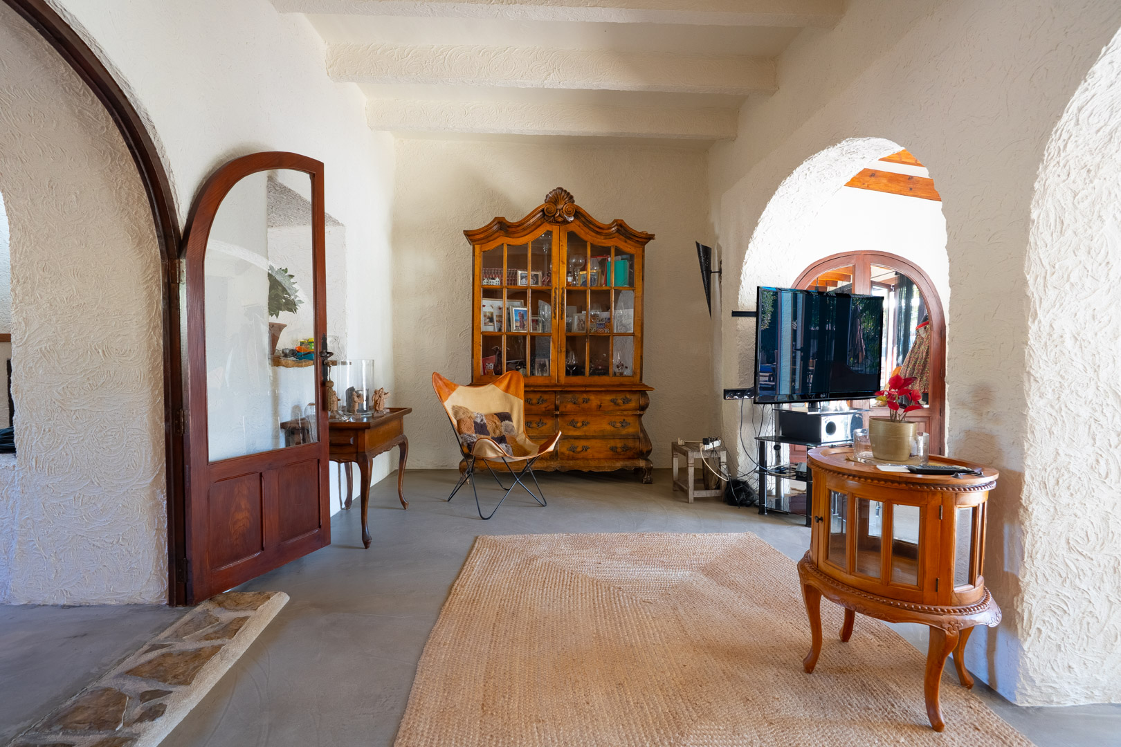 Living area featuring white-beamed ceilings, traditional arches, and microcement flooring. The space includes an antique wooden vitrine and a butterfly chair.