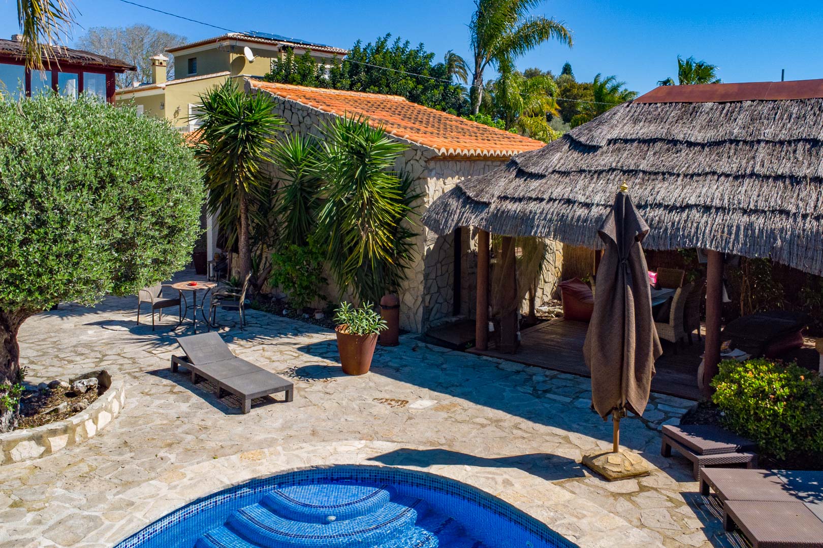 Outdoor area featuring a natural stone patio, swimming pool, and a thatched Balinese pergola adjacent to a rustic stone building and Mediterranean plants.