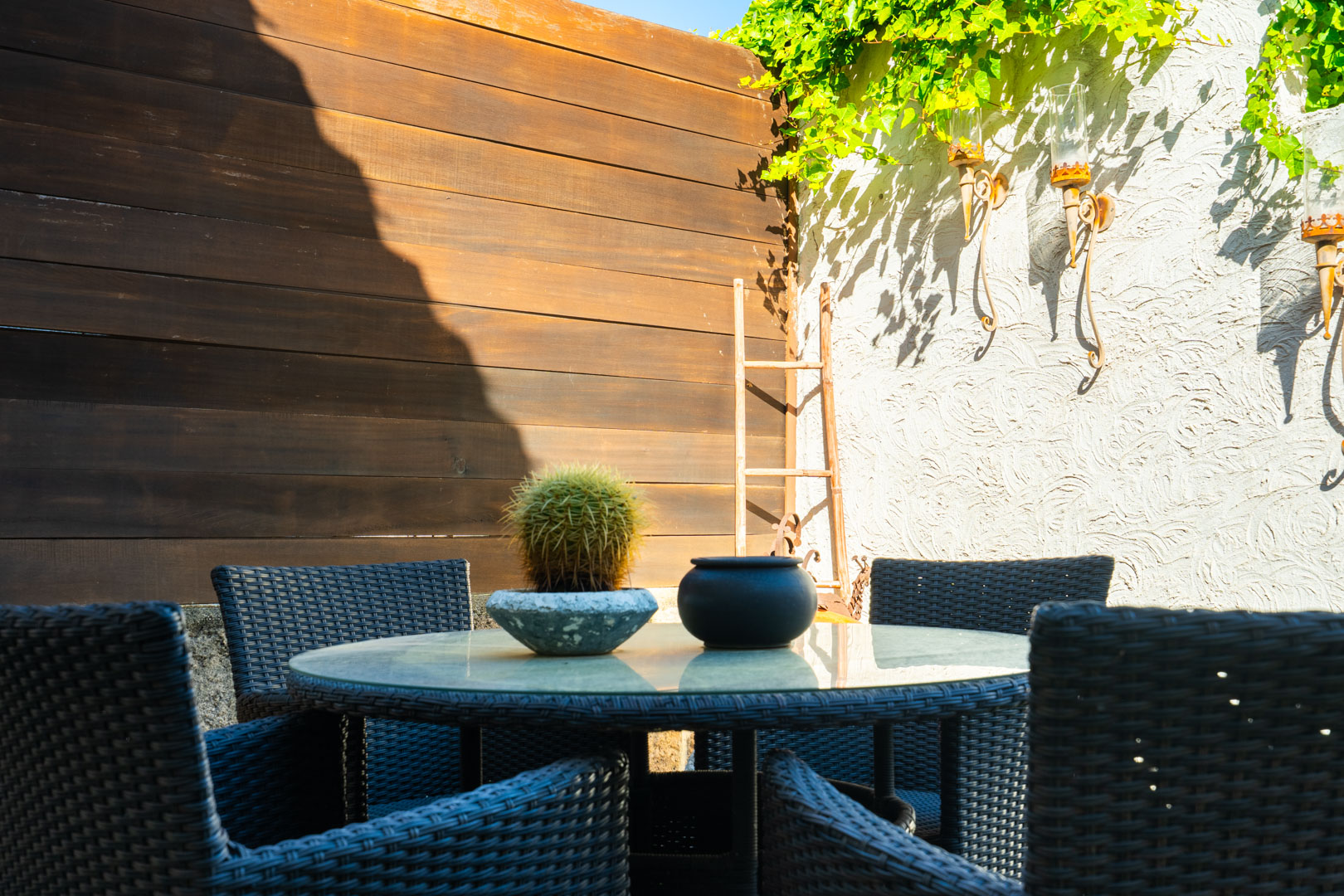 Outdoor dining area featuring a round glass-top table and dark wicker chairs, set against a textured wall with wrought iron sconces and wooden fencing.
