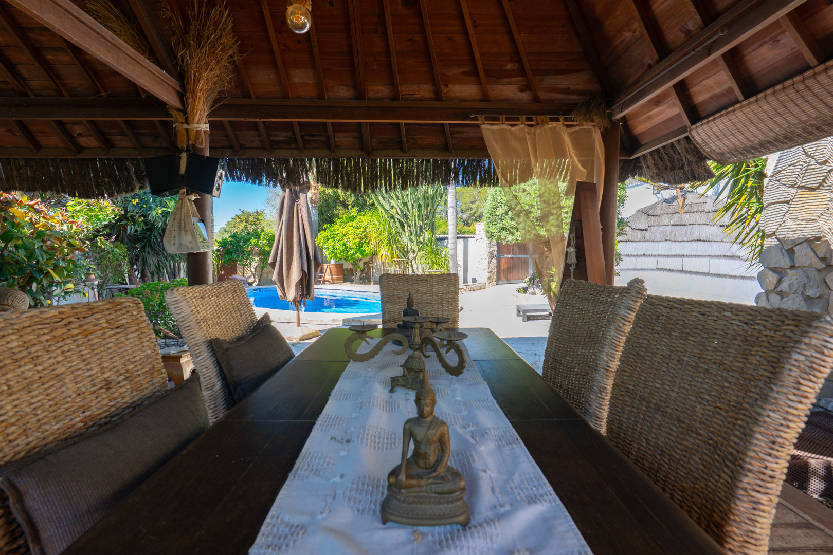 Outdoor dining area under a Balinese wooden pergola, featuring wicker chairs and views of the swimming pool and lush Mediterranean garden.
