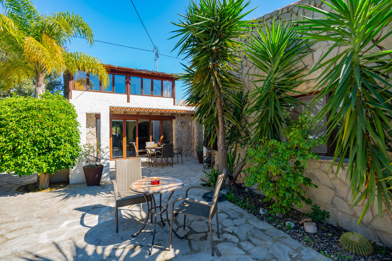 Outdoor patio with natural stone paving, Mediterranean plants, and a facade featuring stone walls and a wooden-framed glazed gallery on the upper floor.