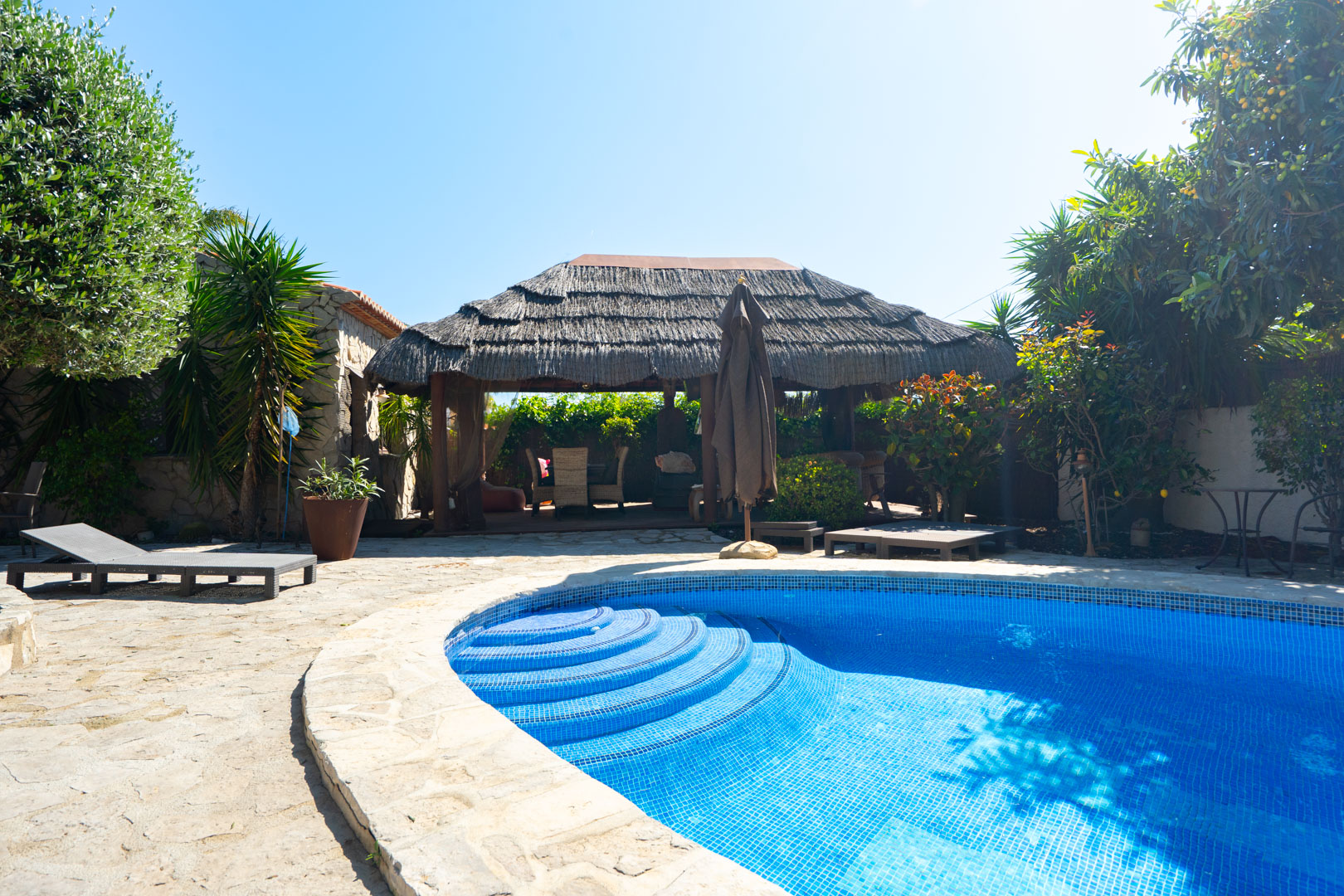 Outdoor swimming pool with blue mosaic tiling and curved steps, featuring a stone terrace and a large thatched Balinese pergola for shaded lounging.