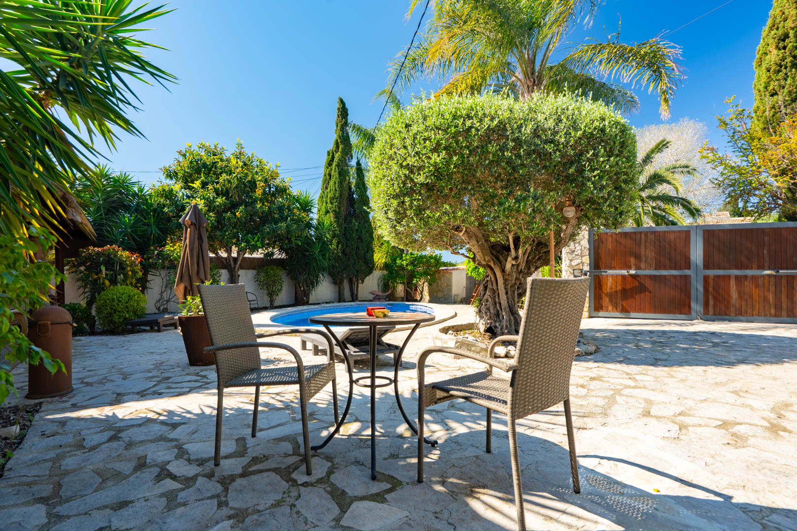 Outdoor terrace featuring natural stone paving, wicker seating, and a view of the private pool framed by mature Mediterranean trees and a wooden entrance gate.