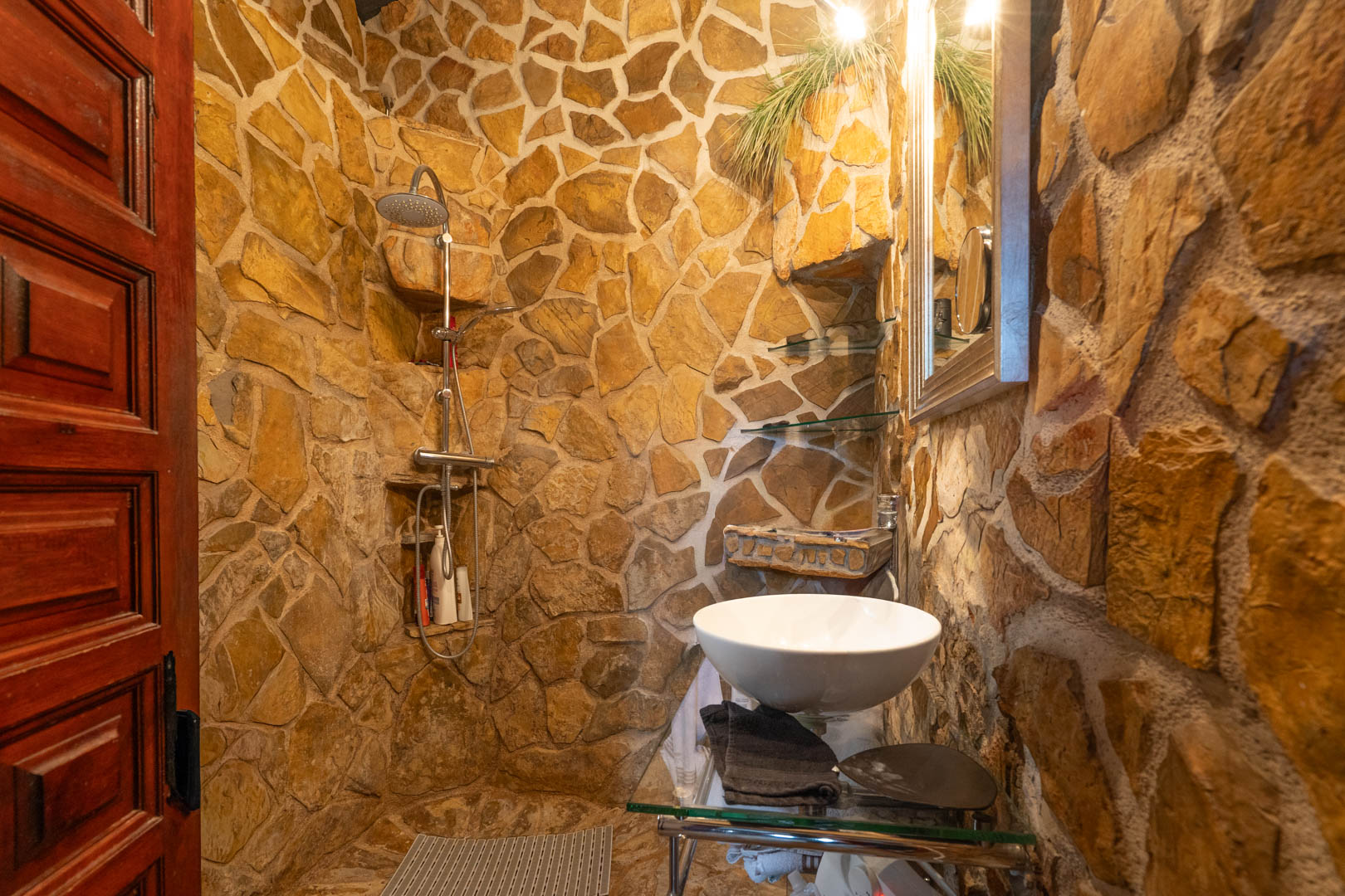 Rustic bathroom featuring natural stone walls, a walk-in shower with integrated shelving, and a vessel sink on a glass vanity.
