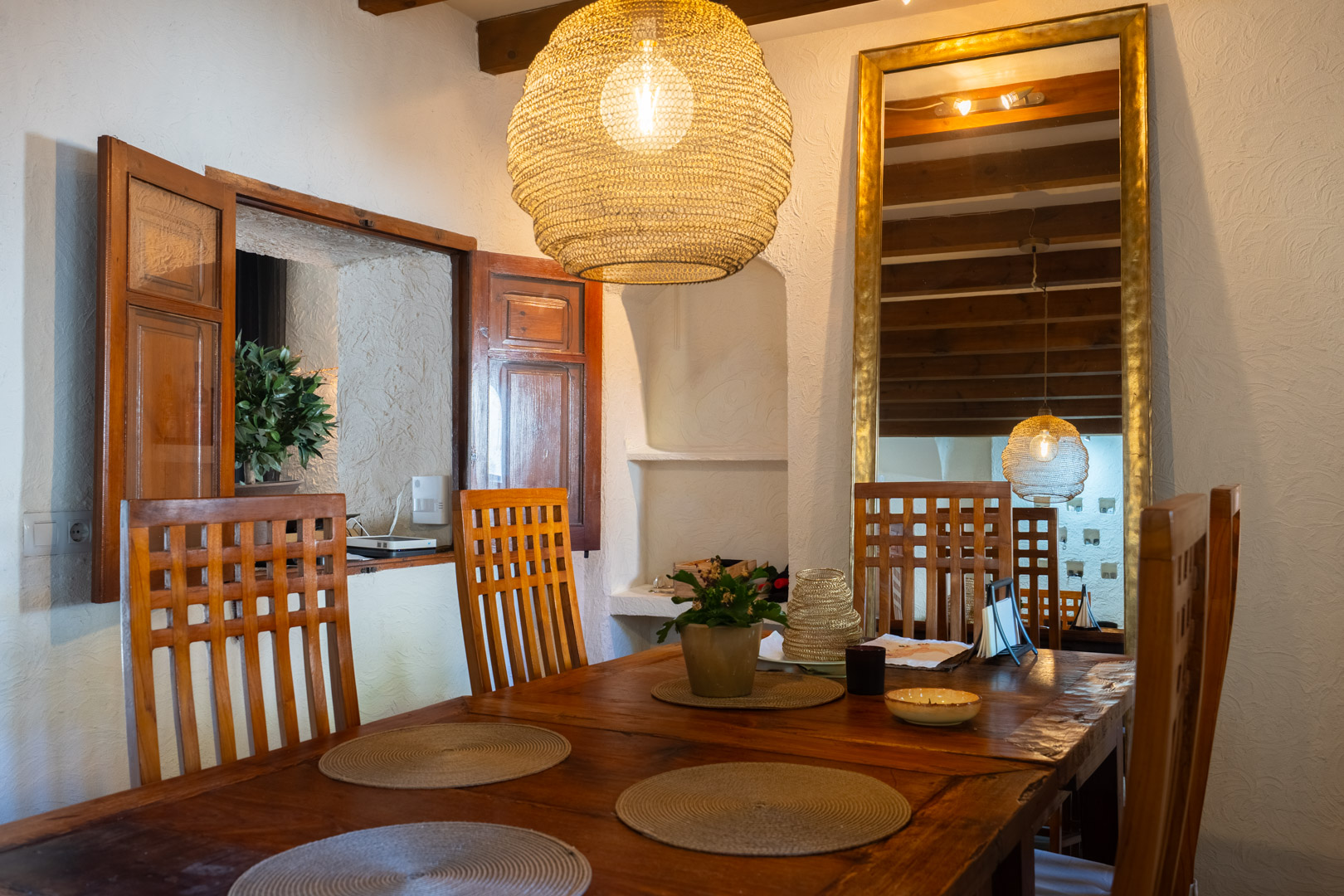 Rustic dining area featuring a solid wood table, high-back chairs, and a natural fiber pendant lamp. The space includes exposed ceiling beams and a large gold-framed mirror.