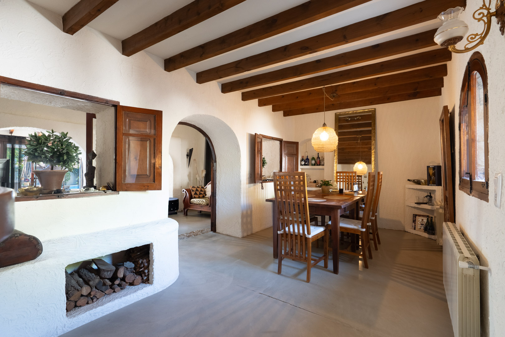 Rustic dining area featuring exposed wooden ceiling beams, a solid wood table, and an arched entryway. Includes a built-in wood storage niche and interior wooden shutters.