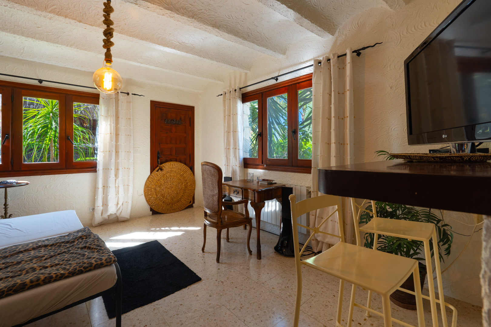 Rustic guest room featuring white beamed ceilings, a classic wooden desk, and dark-framed windows overlooking lush Mediterranean greenery.