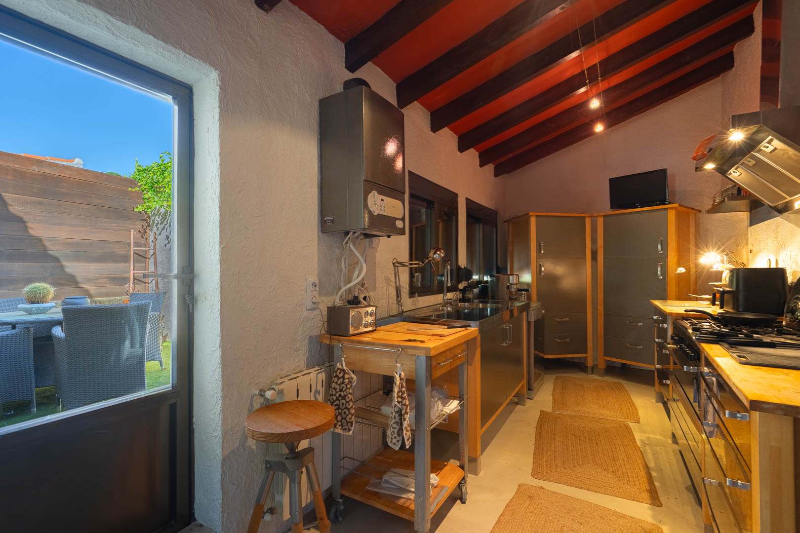 Rustic kitchen featuring exposed dark wood ceiling beams, a mobile butcher block island, and direct access to a furnished outdoor patio.