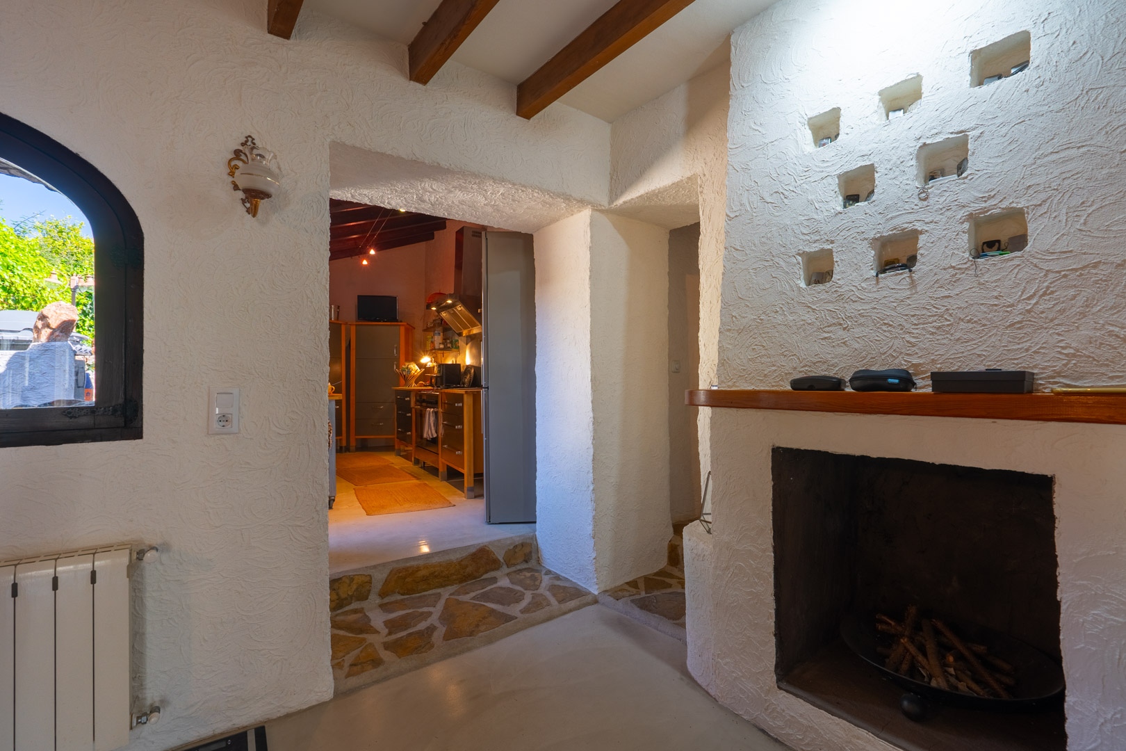Rustic living area featuring exposed wooden beams, a built-in fireplace with decorative niches, and a stone-paved transition leading to the kitchen.