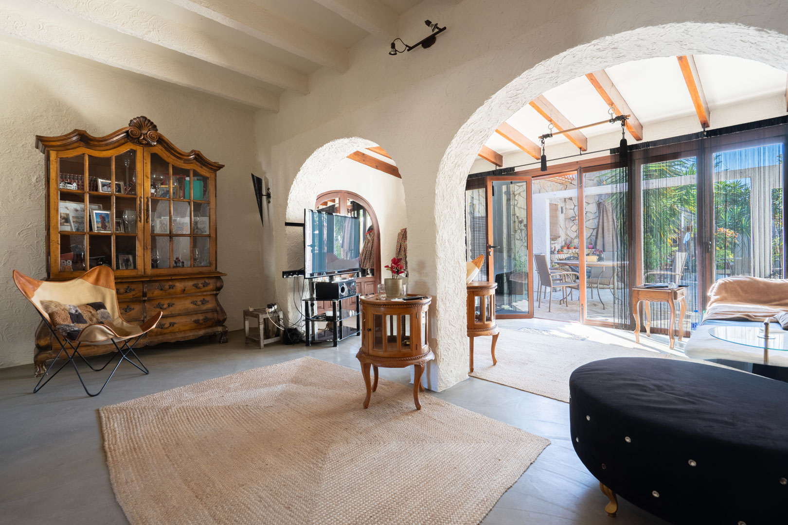Rustic living room featuring white beamed ceilings and arches opening to a naya. Includes an antique wooden cabinet, jute rug, and natural light from the adjacent patio.