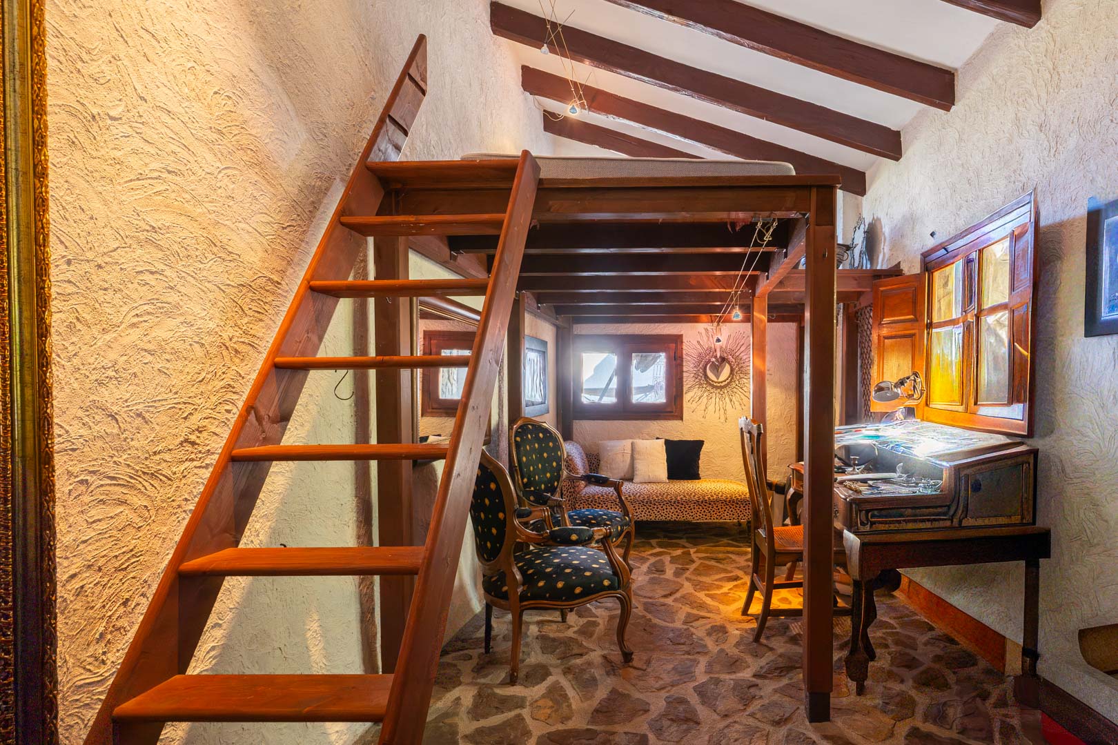Rustic room featuring a wooden mezzanine bed, exposed ceiling beams, and natural stone flooring. The space includes a vintage desk area and eclectic seating.