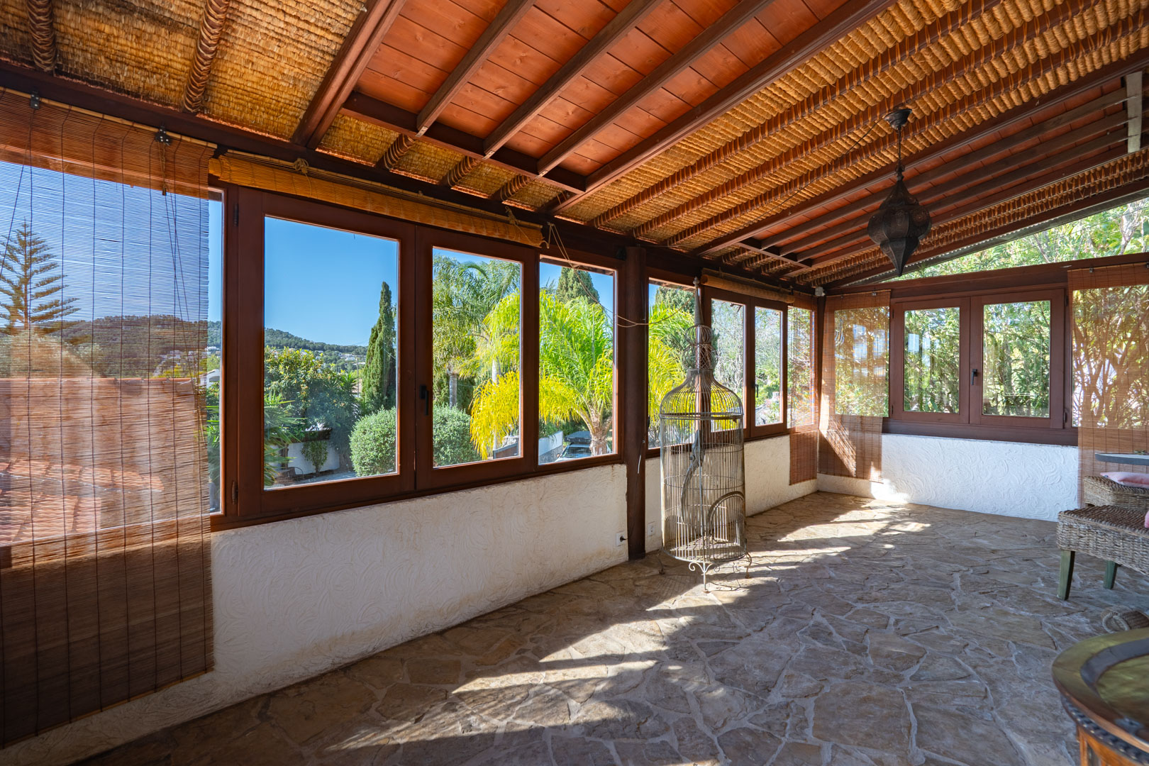 Spacious glazed naya featuring natural stone flooring, exposed wooden beams with reed ceilings, and large windows overlooking the Mediterranean garden.
