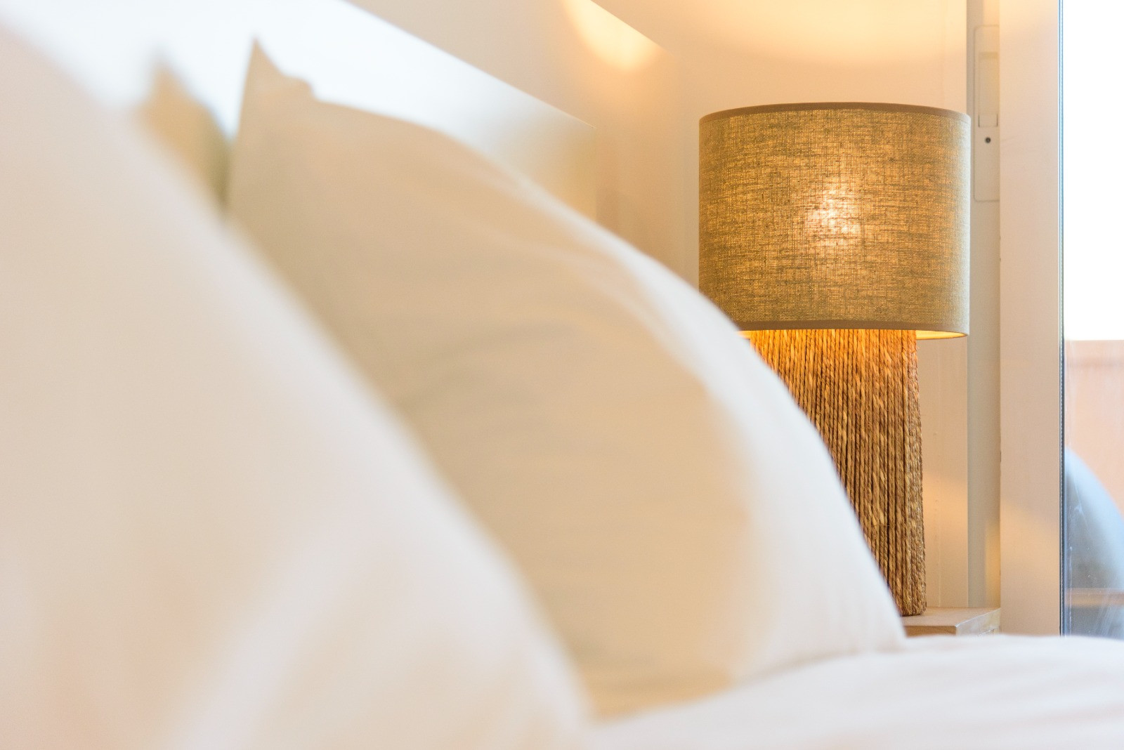 Bedroom detail featuring a bedside lamp with a natural fiber base and linen shade, providing warm ambient light next to crisp white bedding.