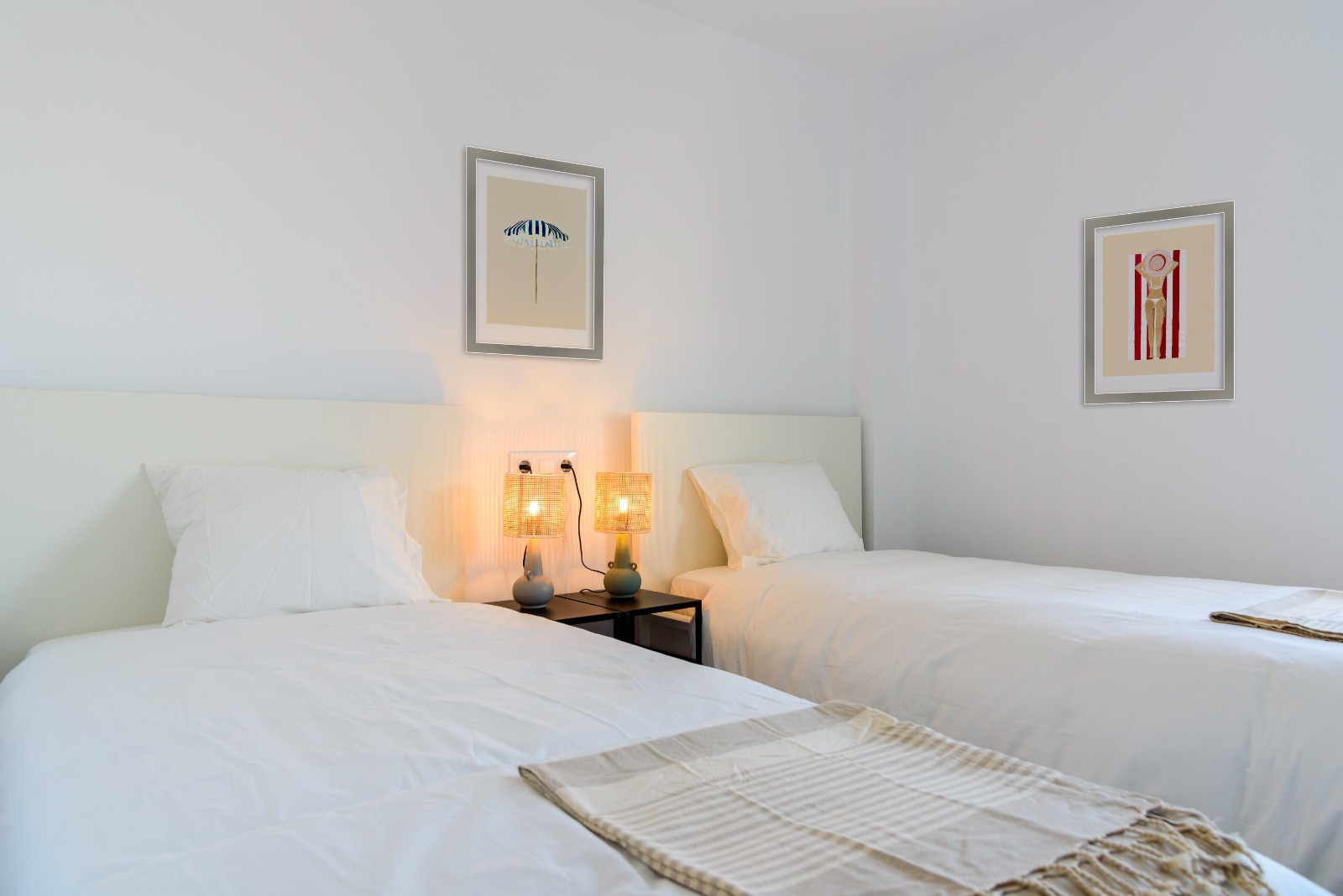 Bright bedroom featuring twin beds with white headboards and natural fiber lamps. The clean white walls are accented by minimalist coastal-themed framed art.