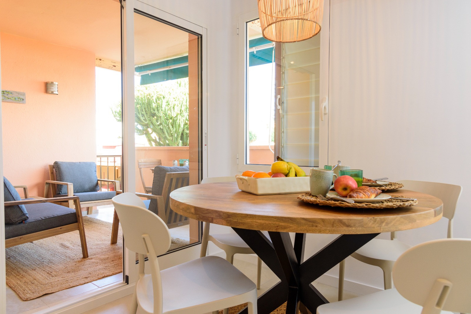 Bright dining area featuring a round wooden table and white chairs, seamlessly connected to a furnished terrace through large glass sliding doors.