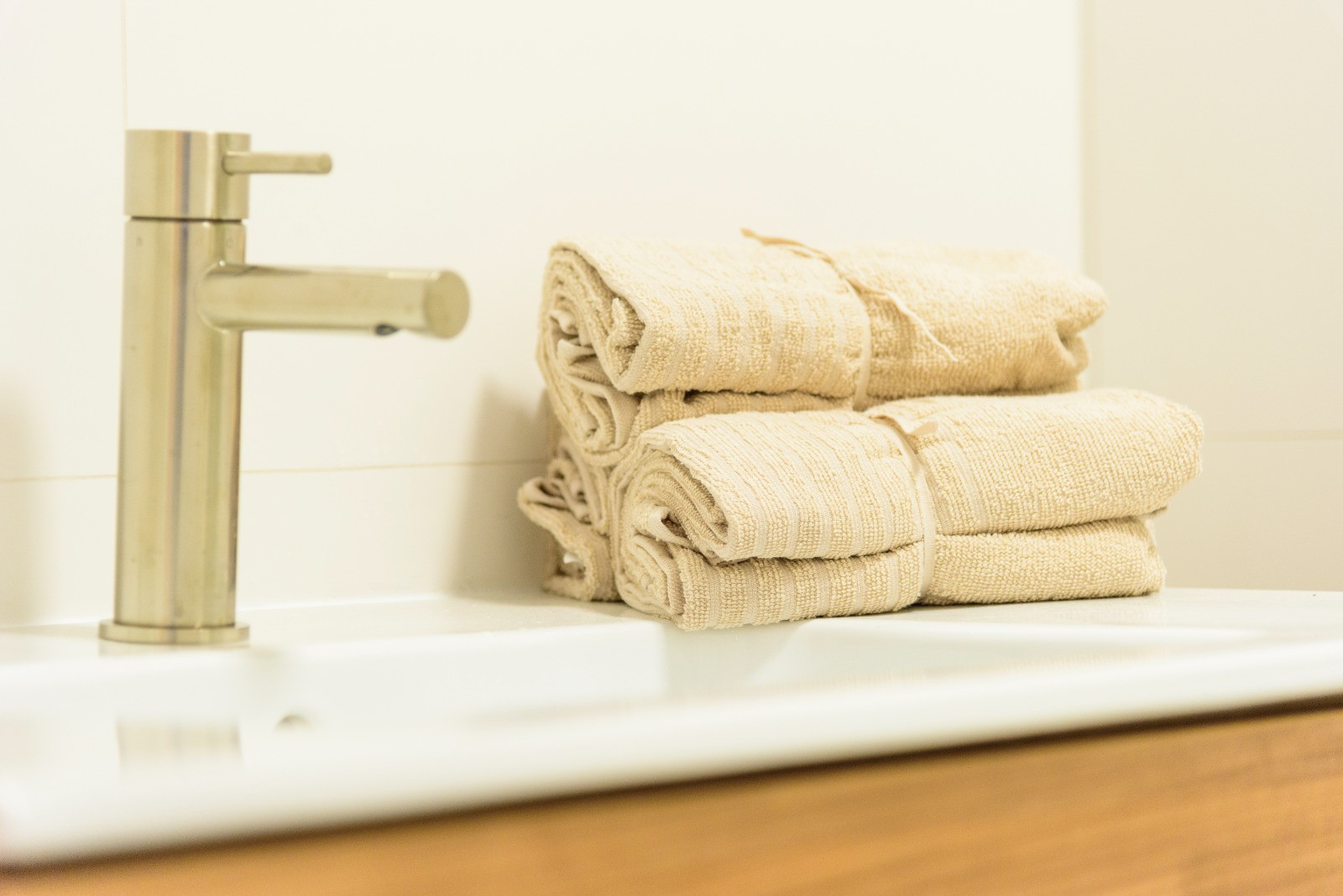 Close-up of the bathroom featuring a brushed steel faucet, white ceramic sink, and neatly rolled beige towels on a modern wooden vanity.