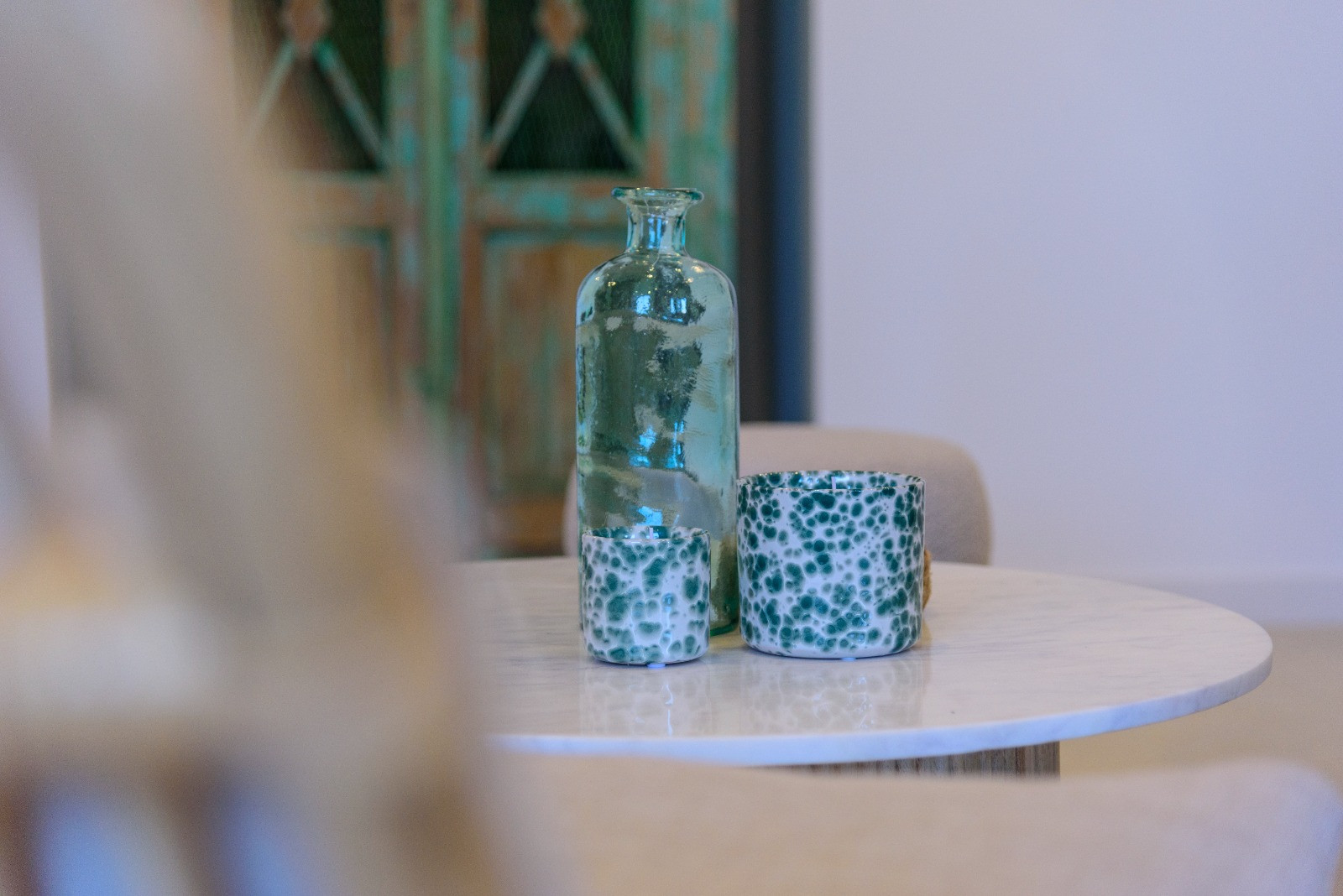 Decorative detail on a white marble table featuring a turquoise glass bottle and patterned ceramic vessels against a blurred rustic background.