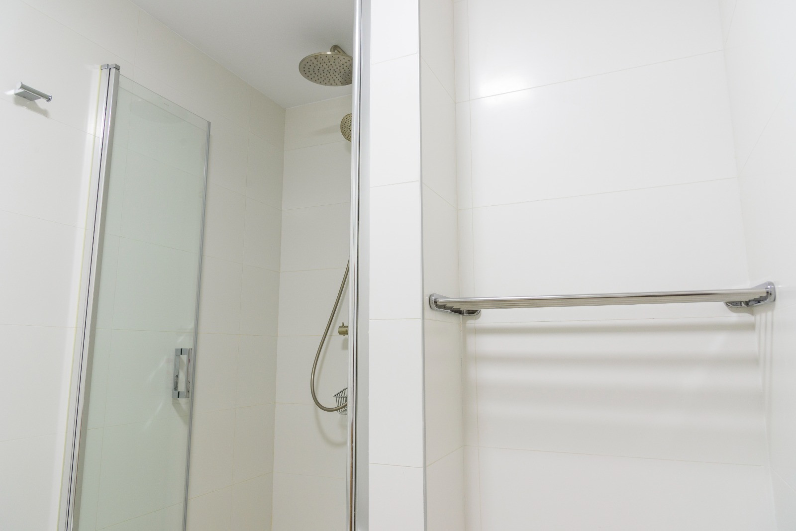 Modern bathroom featuring a walk-in shower with a glass door and chrome fixtures, including a rain shower head. Finished with clean white wall tiles and a metallic towel rack.
