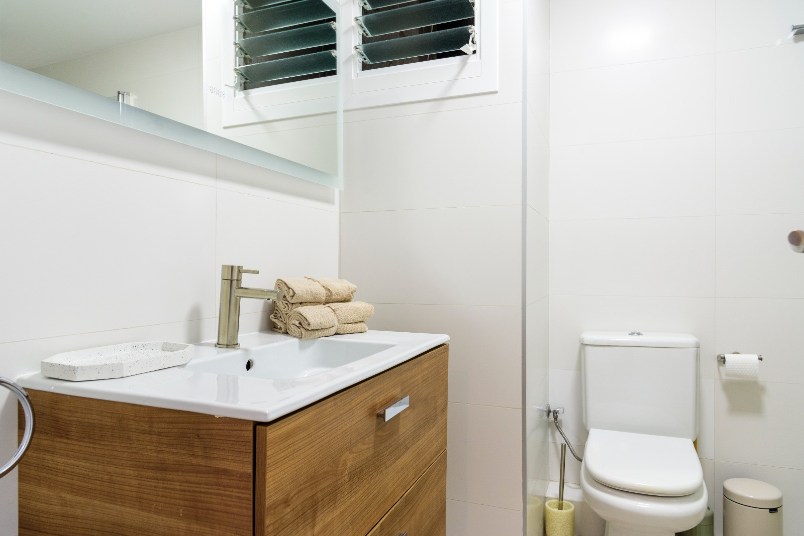Modern bathroom featuring a wooden vanity, integrated sink, and white tiled walls. Includes contemporary fixtures and a louvered window for natural ventilation.