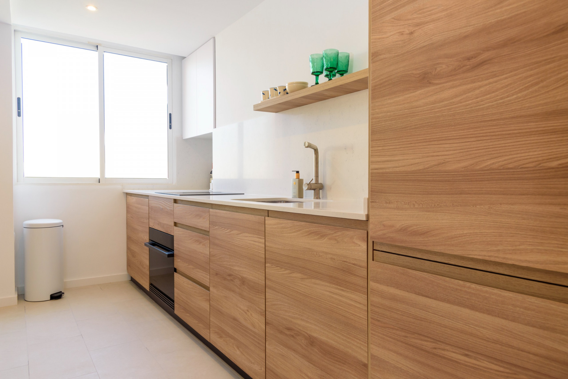 Modern kitchen featuring wood-grain cabinetry, a white countertop, and an integrated sink. A large window fills the space with natural light, highlighting the clean, functional design.