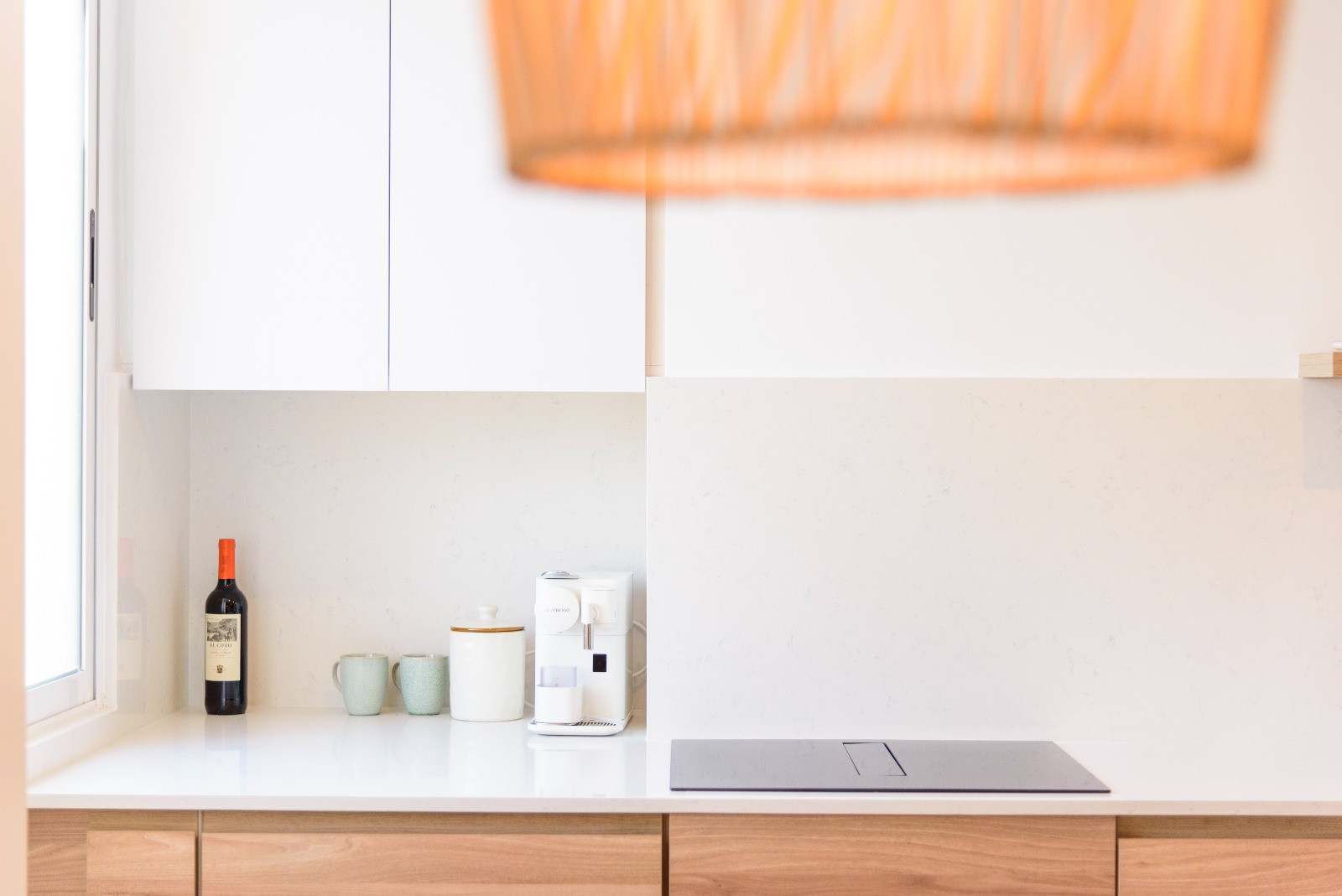 Modern kitchen with white stone countertops, integrated induction hob, and wooden lower cabinets. Natural light from the window highlights the minimalist coffee station and clean finishes.