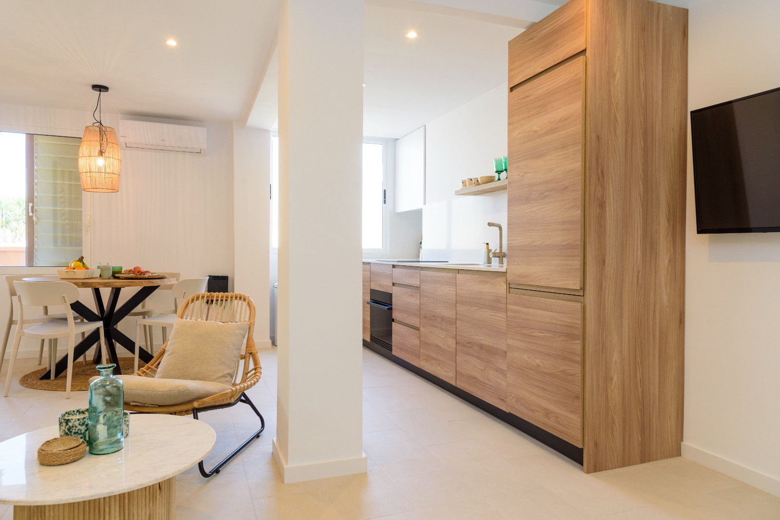 Open-plan living space featuring a modern wood-finish kitchen, dining area with a rattan pendant light, and neutral porcelain flooring.