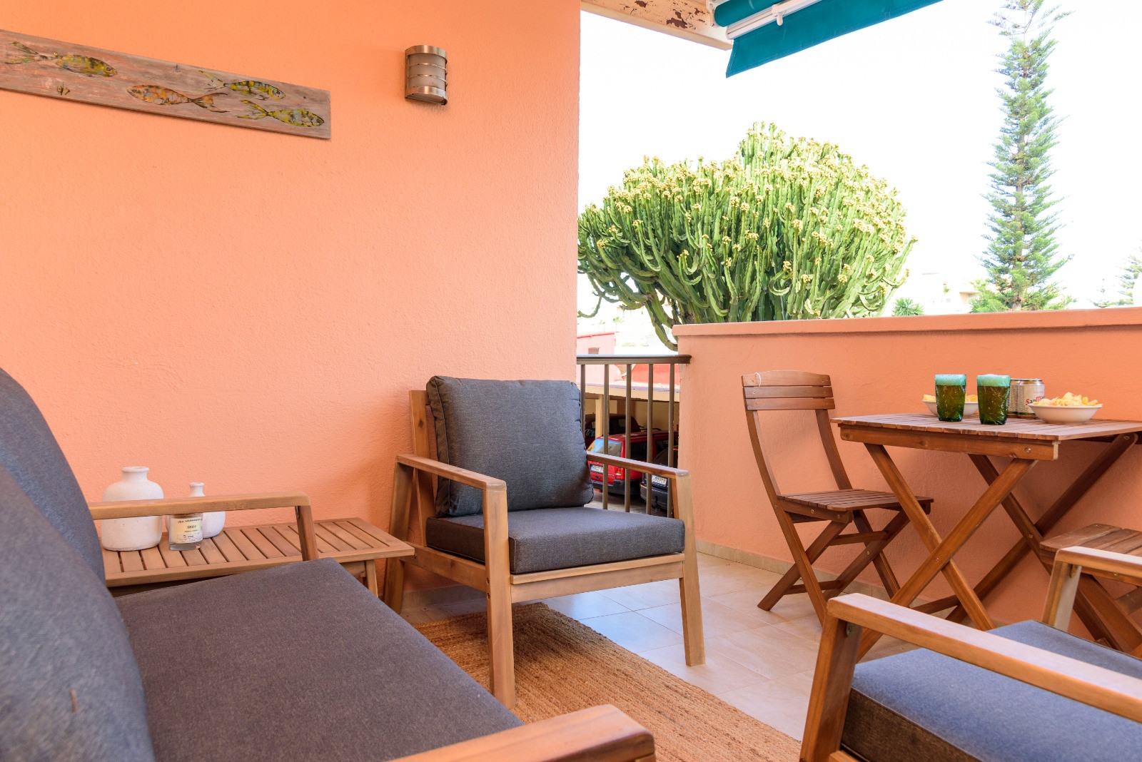 Private terrace featuring wooden lounge furniture with grey cushions, an outdoor dining set, and views of local greenery under a retractable blue awning.