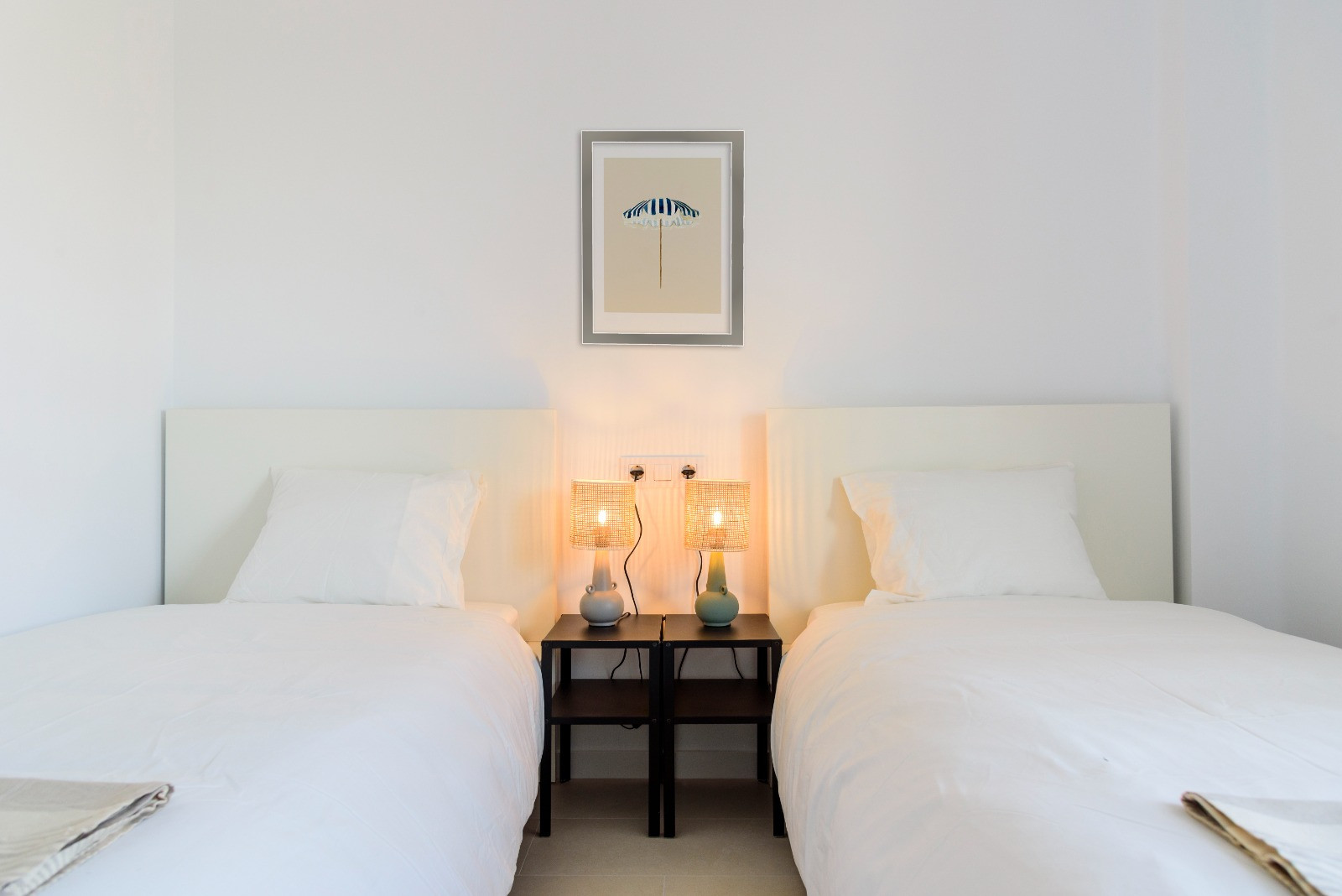 Twin bedroom featuring white headboards and central black nightstands with woven lamps, complemented by a minimalist beach-themed artwork on white walls.