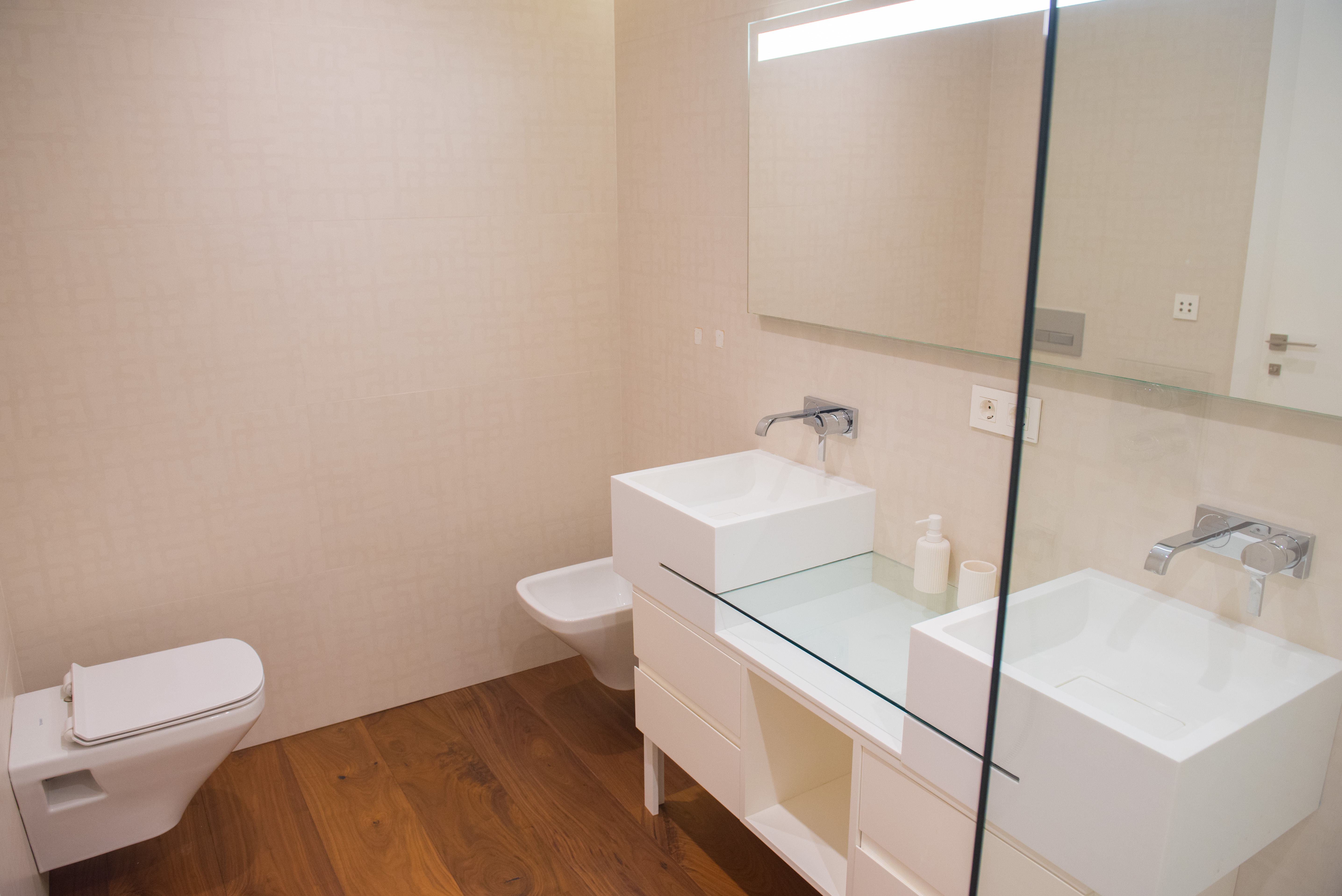 Contemporary bathroom with double square vessel sinks, glass countertop, and wall-mounted faucets. Features wall-hung sanitary ware and warm wood flooring.