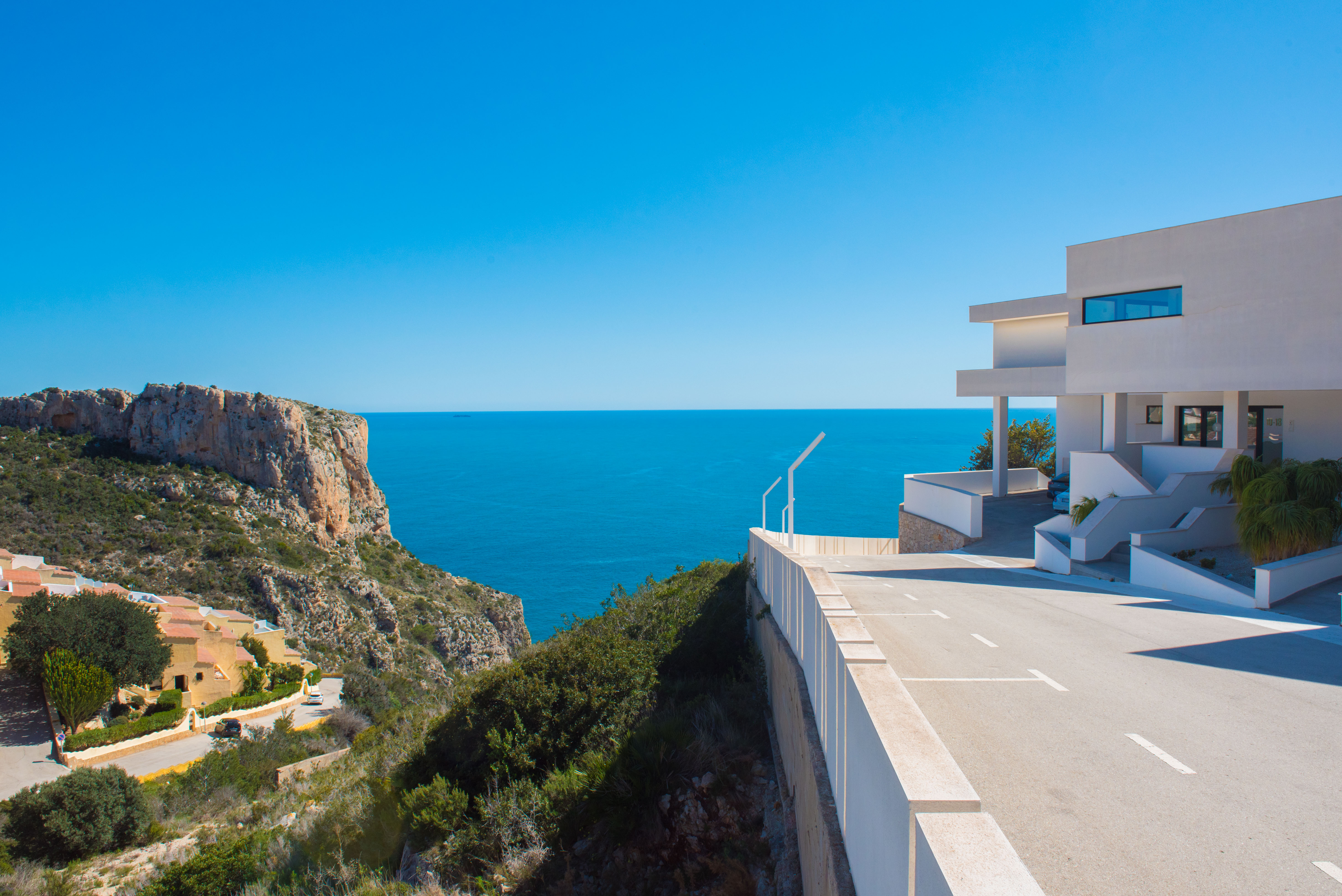 Exterior view of the minimalist white building complex on a cliffside, featuring a paved access area and expansive views of the Mediterranean horizon.