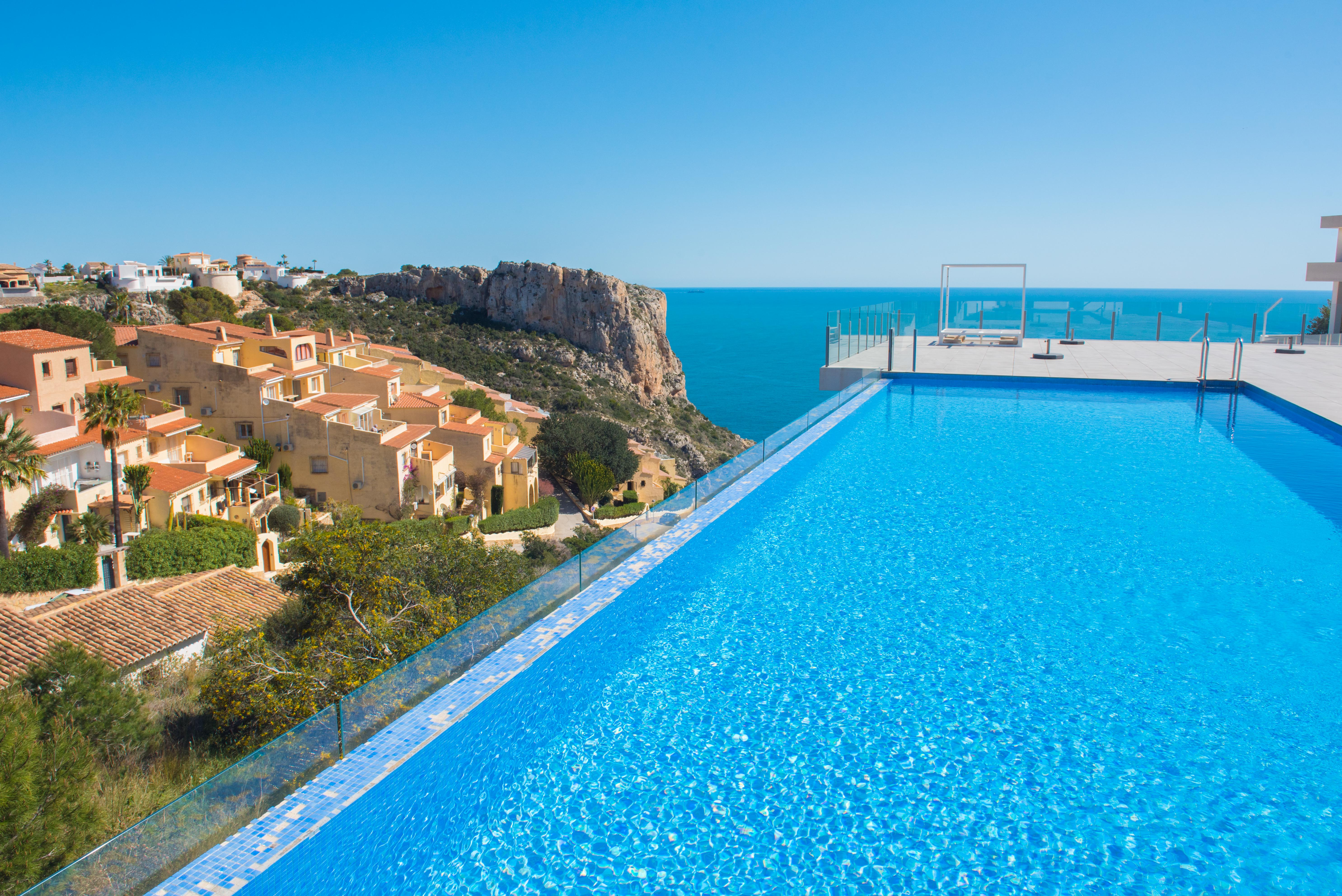Infinity pool featuring blue mosaic tiles and glass railings, overlooking the Mediterranean Sea, coastal cliffs, and Mediterranean-style villas.