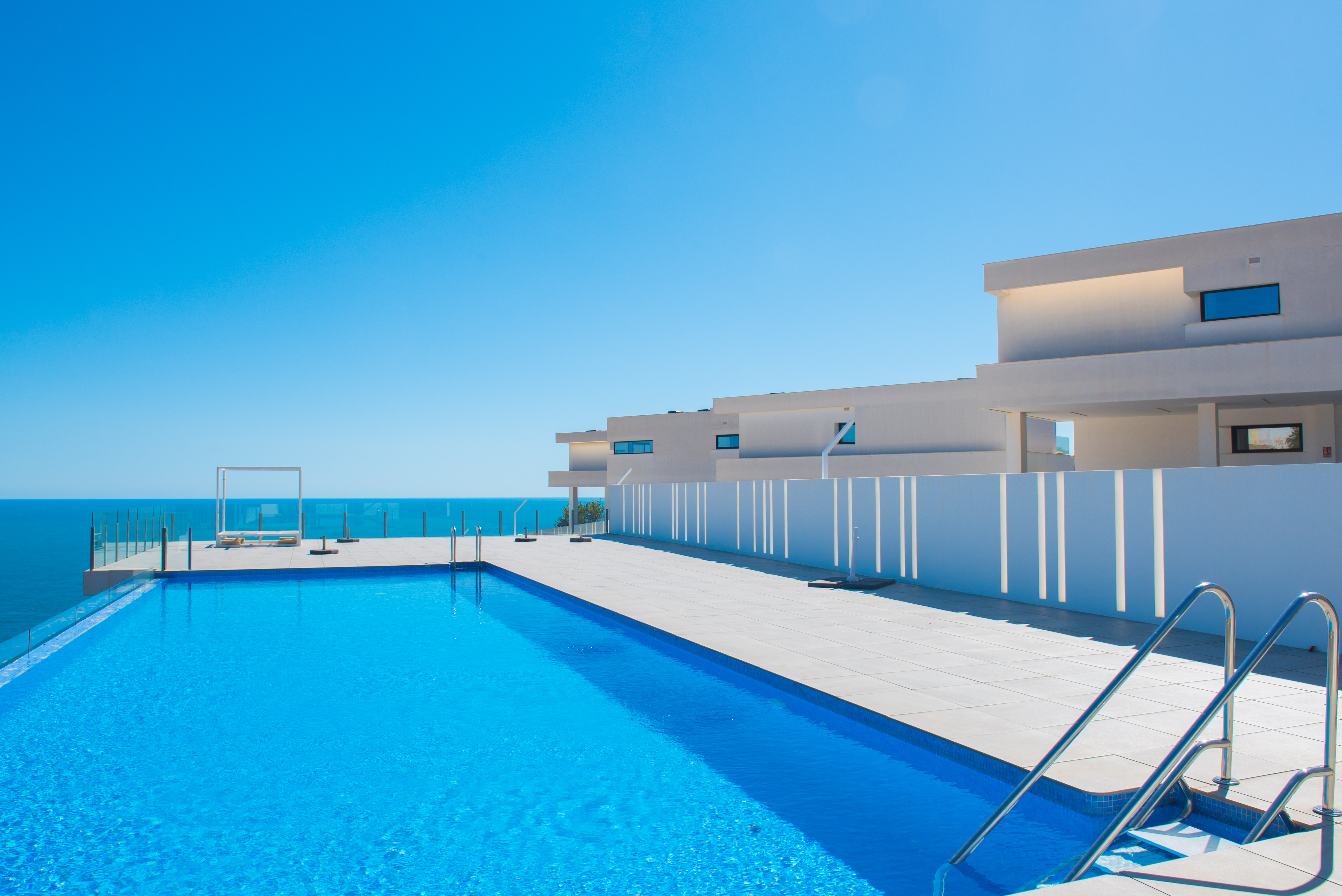 Infinity pool with Mediterranean views, featuring a spacious tiled terrace, glass railings, and minimalist white architecture under a clear sky.