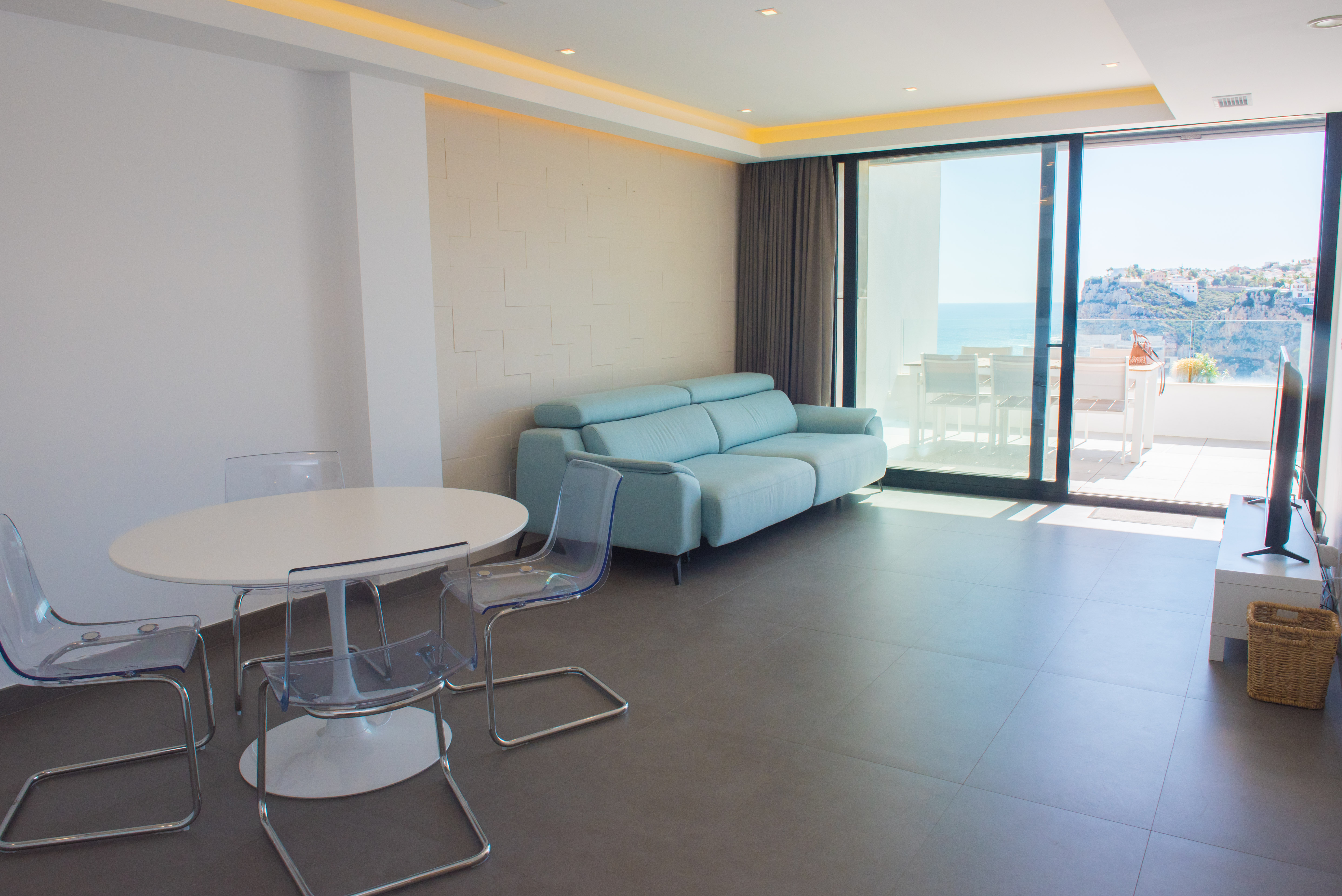 Living-dining space featuring a pastel blue sofa, acrylic chairs, and recessed ceiling lighting. Large sliding doors connect the interior to a terrace with sea views.