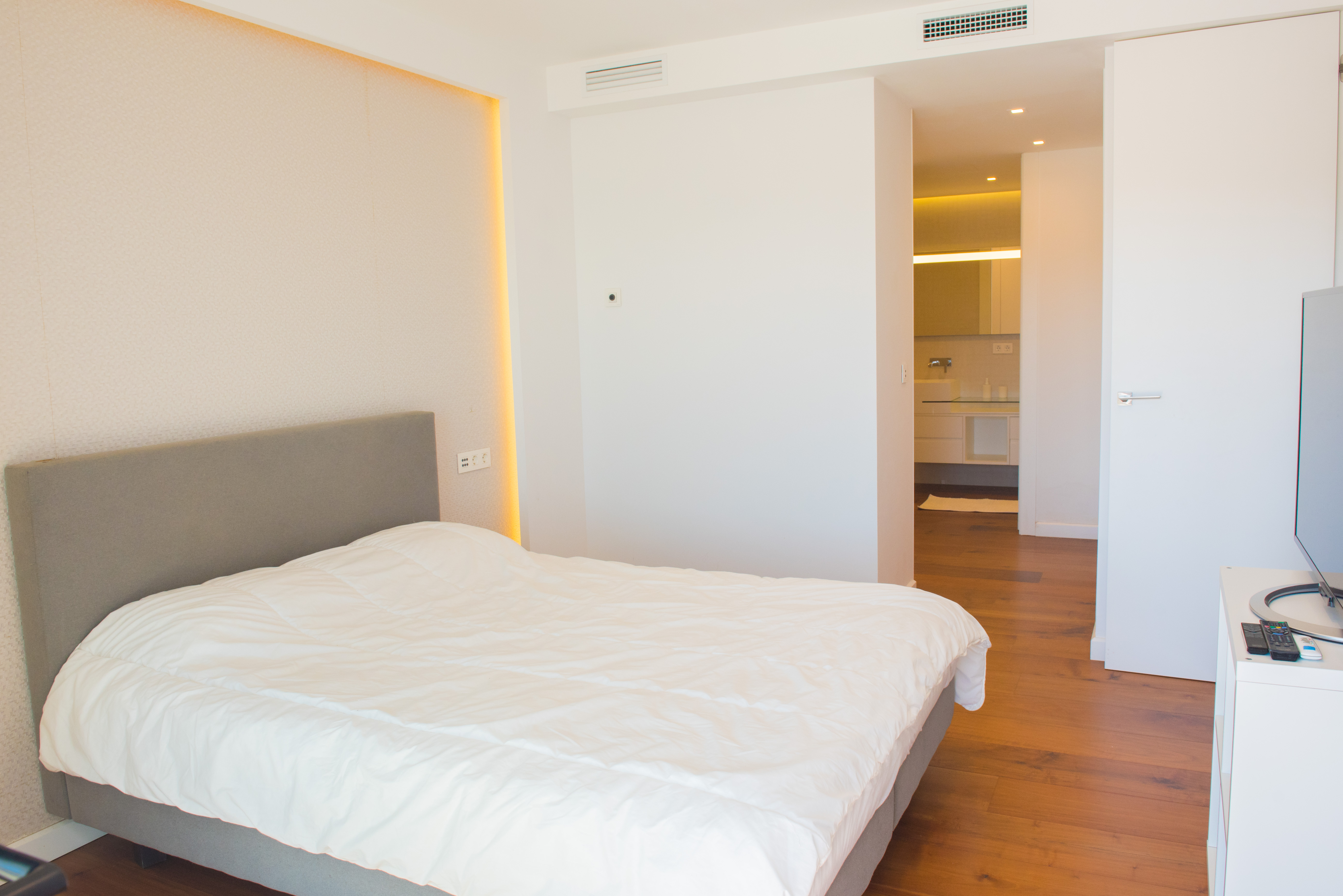 Master bedroom featuring a grey upholstered headboard, indirect LED wall lighting, warm wooden flooring, and a glimpse into the modern en-suite bathroom.