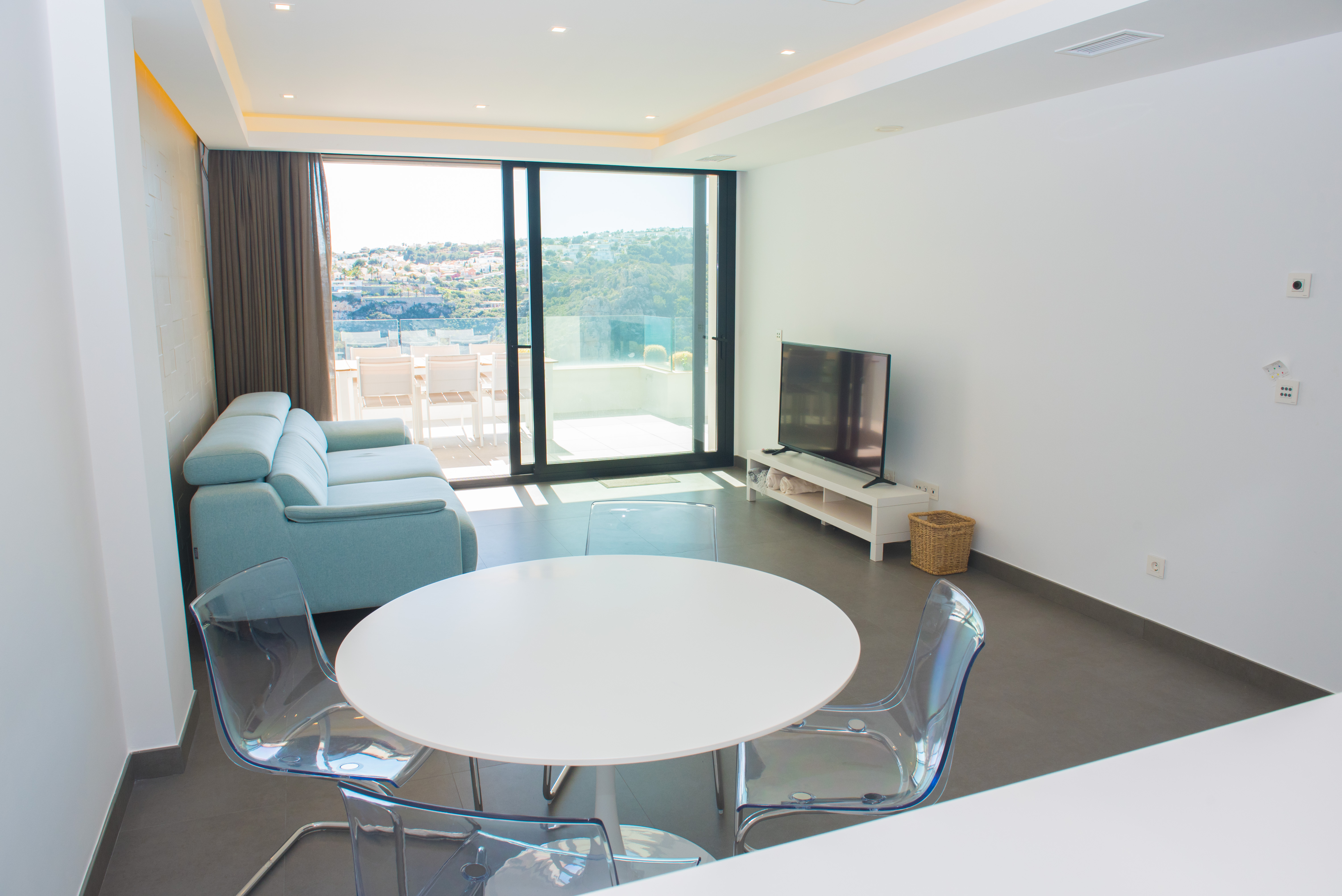 Minimalist living-dining area with a white circular table, acrylic chairs, and light blue sofa, featuring large sliding glass doors that lead to a scenic terrace.