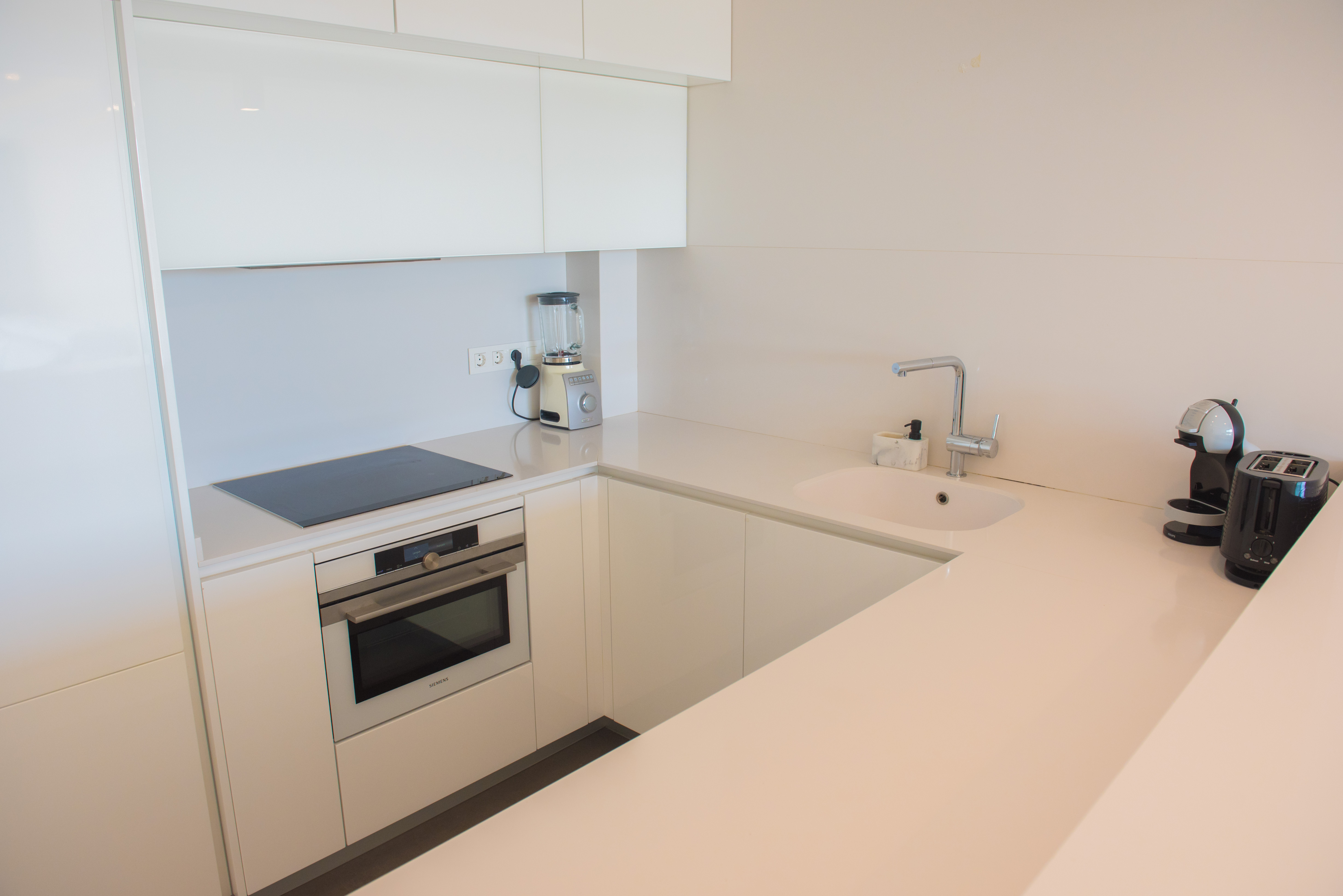 Minimalist white gloss kitchen featuring quartz countertops, a Siemens oven, induction cooktop, and premium small appliances in a functional U-shaped layout.