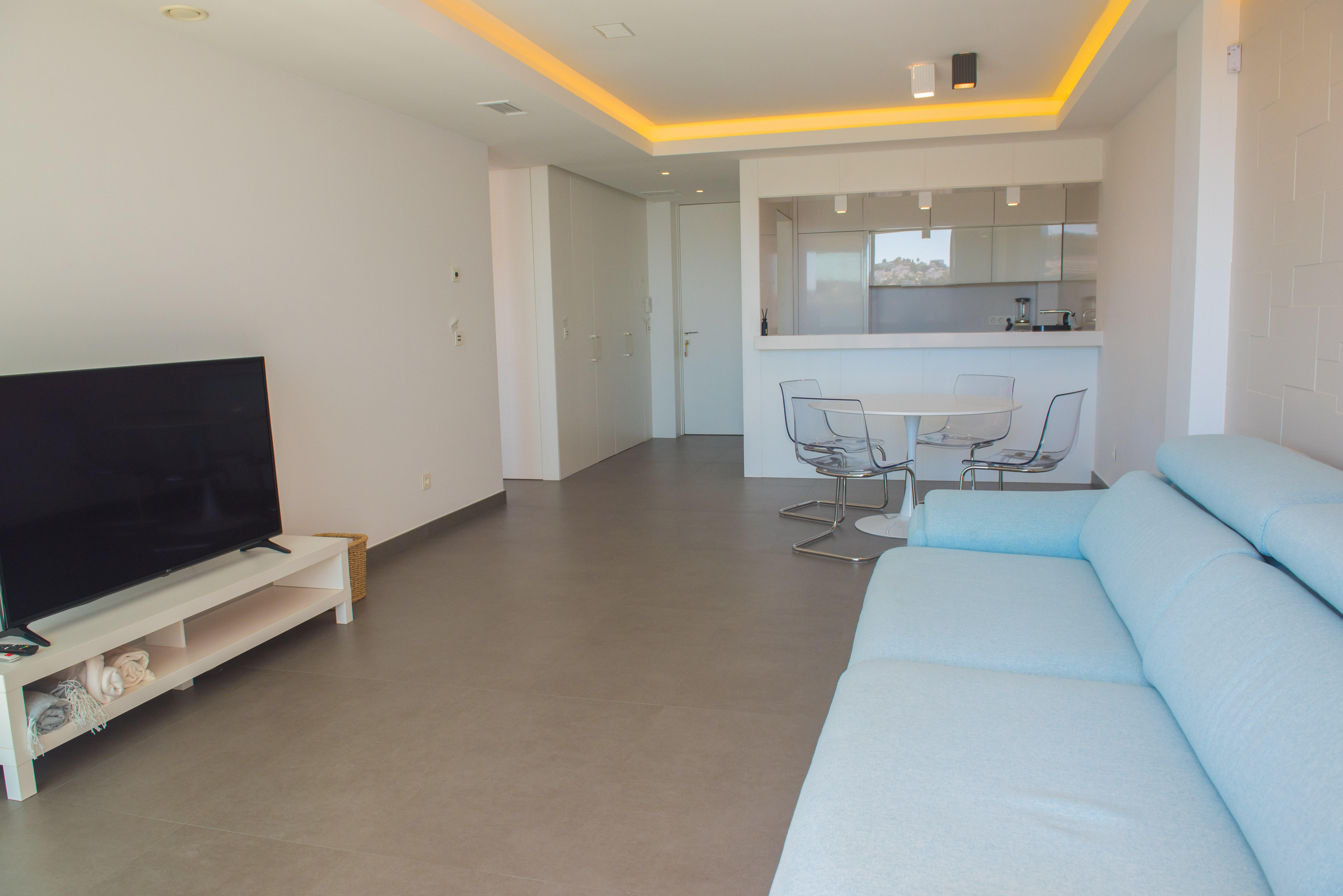 Open-concept living room featuring a light blue sofa, transparent dining chairs, and an integrated kitchen with recessed perimeter lighting.